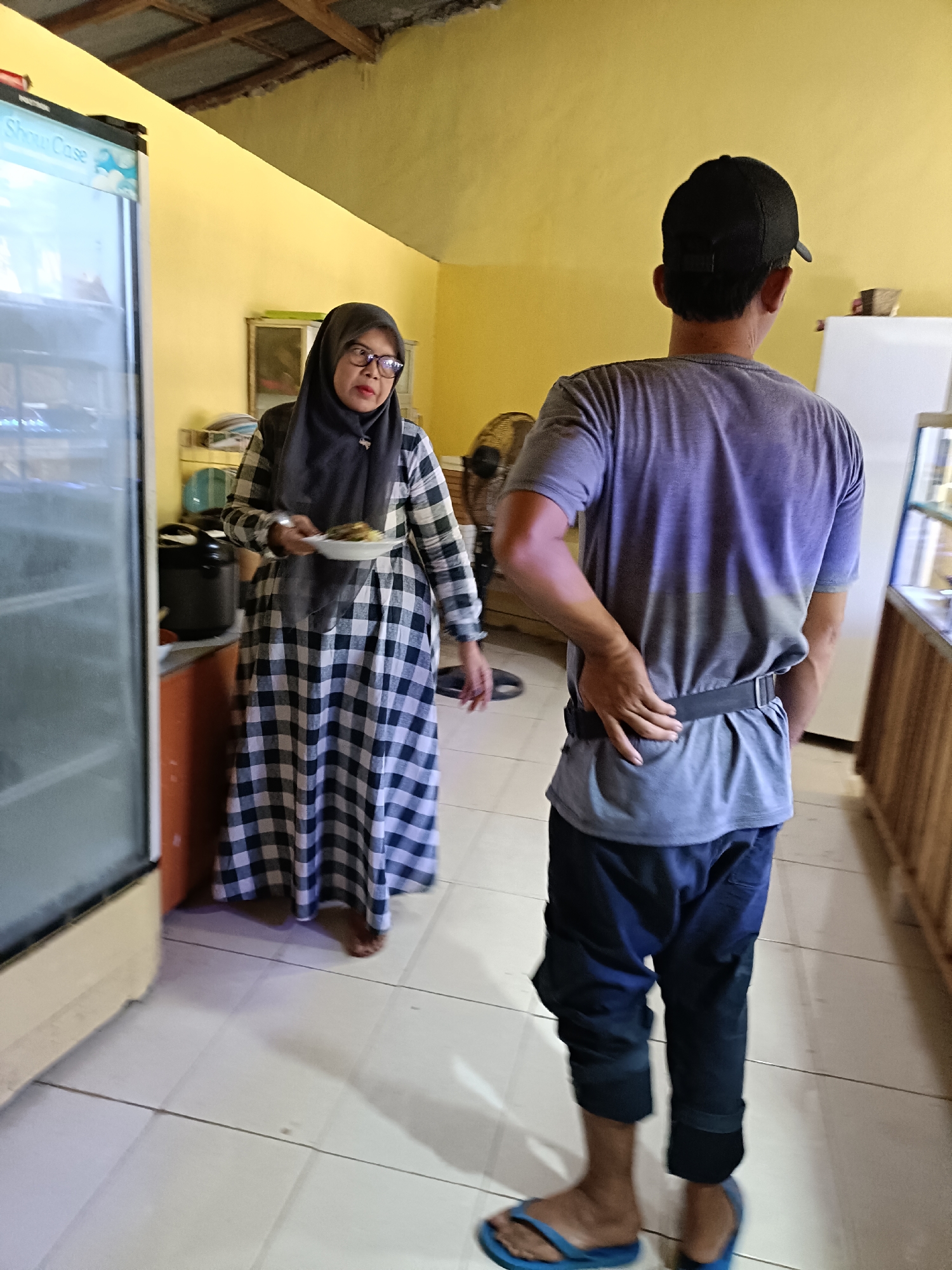 In a brightly lit room with yellow walls and a corrugated metal ceiling, a woman wearing a grey and black checkered dress and a hijab holds a plate of food. She is standing next to a glass-fronted refrigerator with "Show Case" written on it. To her right, a man wearing a grey t-shirt, dark pants, and a black baseball cap stands with his back to the camera, one hand resting on his lower back. A standing fan is visible behind him, and a counter with items on it is to his right. The floor is tiled with off-white square tiles. The scene appears to be indoors, possibly in a small shop or eatery in Pordapor, Indonesia.