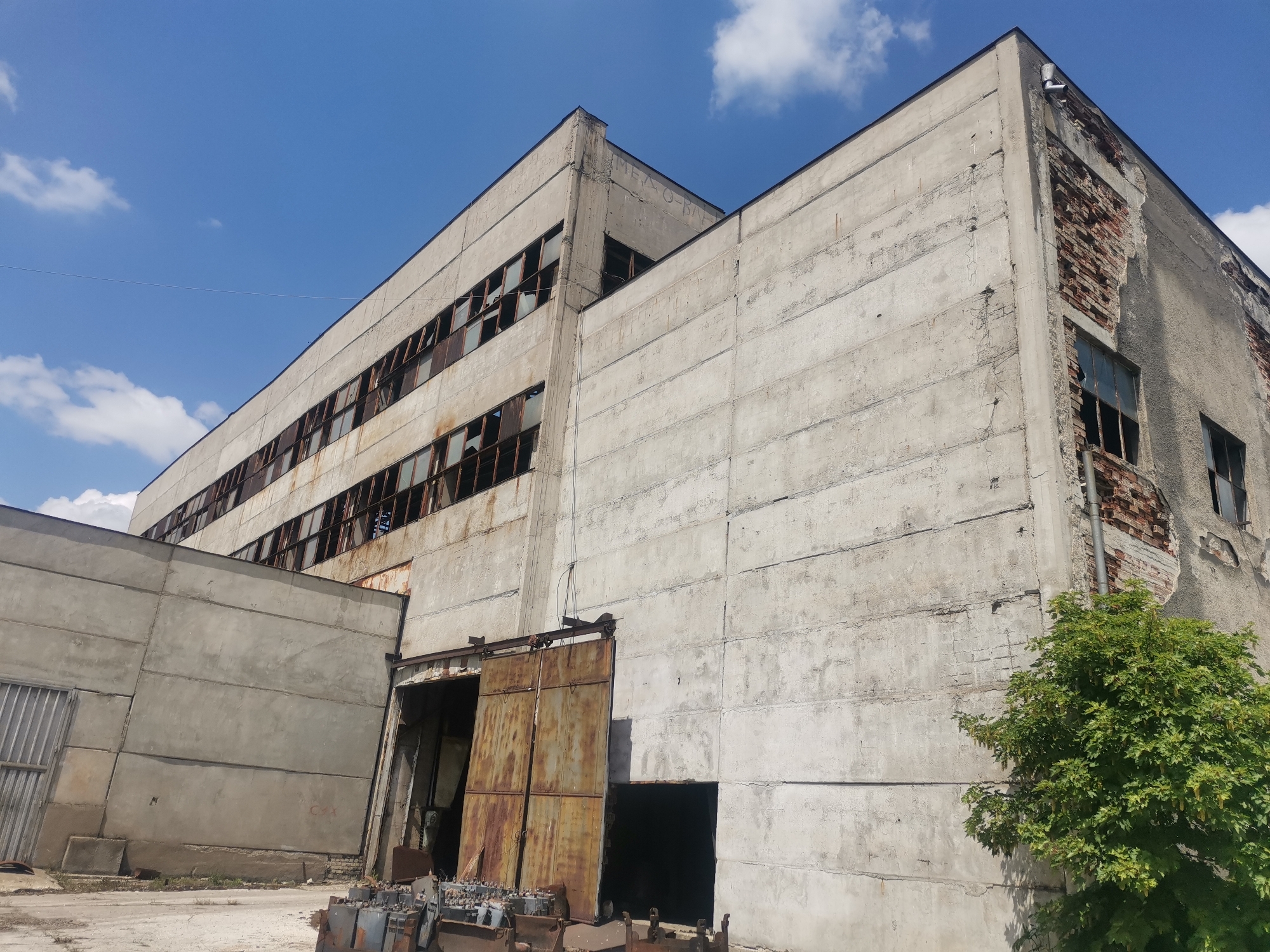The image depicts an abandoned, dilapidated industrial building in what is claimed to be Zhivkovo, Bulgaria. The structure is predominantly constructed of concrete panels, many of which are stained and show signs of age and weathering. A section of the building reveals exposed brickwork where the facade has crumbled. Numerous windows are broken and missing panes, with rusting frames. A large rusty metal sliding door is partially open, revealing a dark interior. The ground is littered with discarded metal objects and debris. A small tree grows near the building. The sky is visible, showing a clear blue color with scattered white clouds.