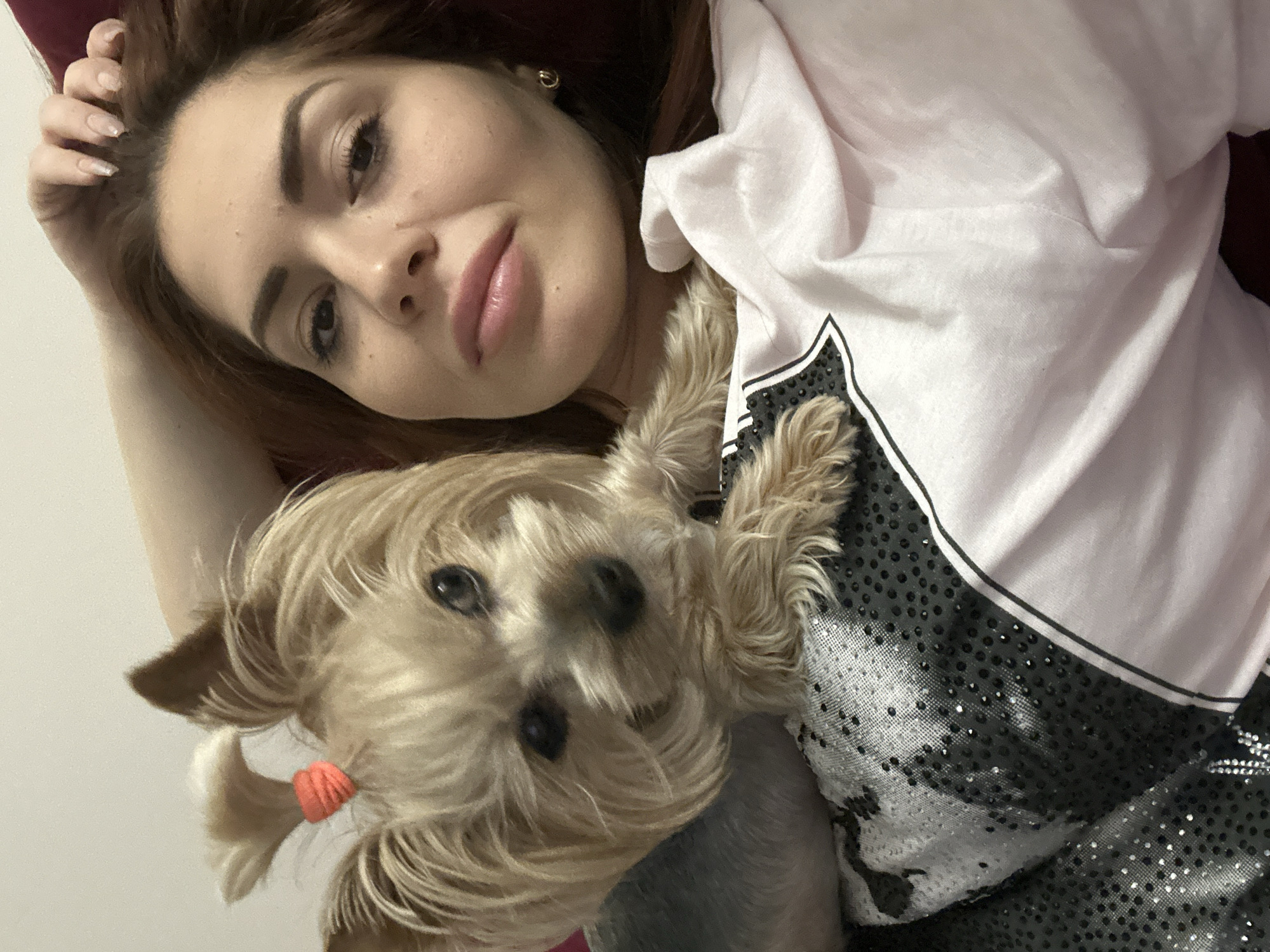 A woman and a dog are in the foreground of the image. The woman is fair-skinned with auburn hair and is wearing a light-colored top with a black and silver design. She is lying down and looking at the camera. Next to her is a small tan-colored dog with its hair pulled into a small ponytail secured with a pink rubber band. The background is a neutral color. The location is not discernible from the image.