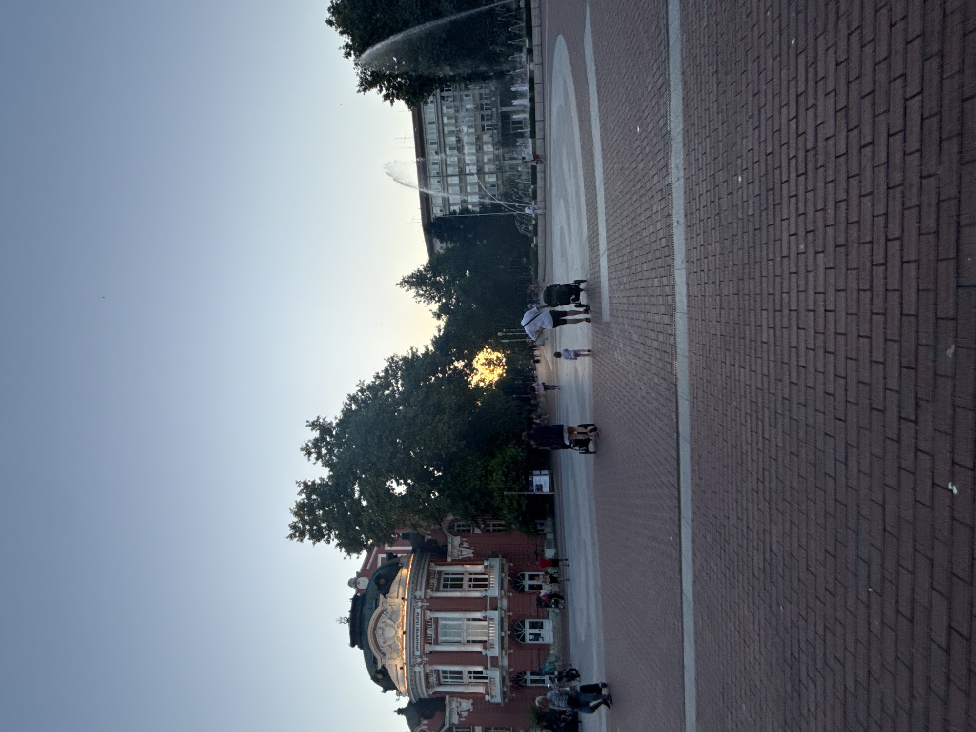 In an outdoor urban plaza at dusk, a red and white ornate building with a domed roof stands to the left, displaying text that appears to be "ДРАМАТИЧЕН ТЕАТЪР" in Cyrillic. In the background, a large multi-story building with many windows is visible. A prominent fountain with several streams of water erupting into the air is in front of this building. The plaza's ground is paved with red bricks. Several people are scattered throughout the scene. In the foreground, a person pushes a stroller, and a small child walks beside them. To the left of the main building, a woman with a stroller is walking. Further to the left, near the ornate building, two people on a scooter are visible. More individuals and strollers are present further back in the plaza, and trees line the perimeter. The sky is a clear, pale blue, indicating the end of daylight.