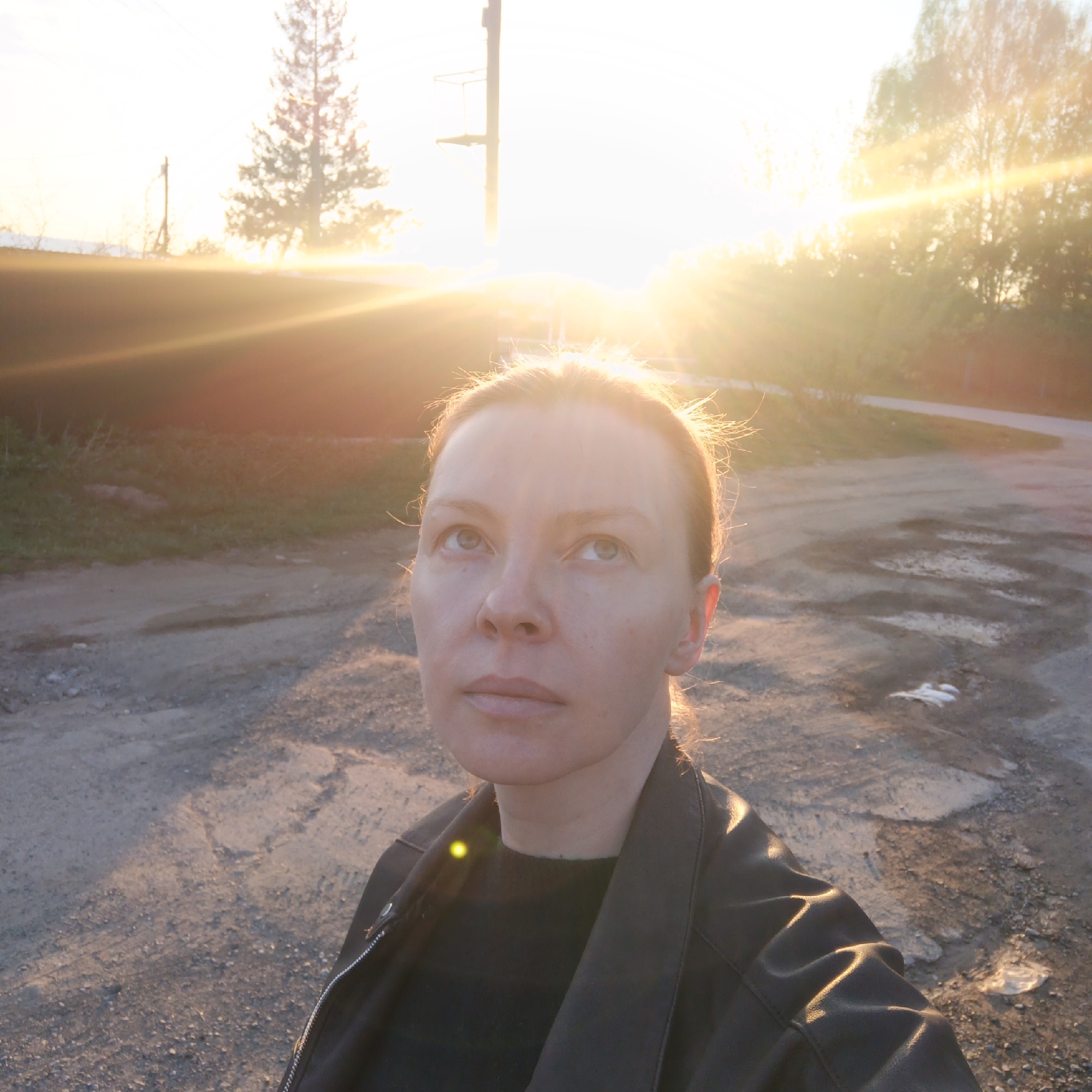 A selfie taken at sunset in what appears to be Chaikovsky, Russia, as the surrounding terrain matches the area. A light-skinned woman is the focal point of the image, wearing a black leather jacket over a dark top. She gazes upwards and slightly to the left. The sun is setting behind her, creating a lens flare that washes out the background detail. Behind the subject, a barren landscape consisting of a dirt road and some vegetation is visible. An electric pole and a pine tree are backlit by the setting sun in the background. The road surface has visible marks and minor puddles. The light suggests that the photo was taken either early morning or late afternoon.