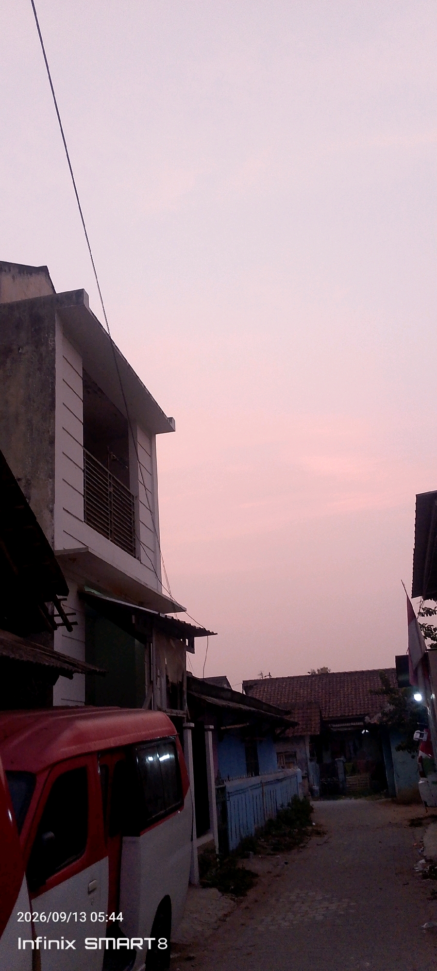 A street scene in Serang, Indonesia, is captured at dusk with a pale pink sky. In the foreground, a red and white van is parked, partially obscuring a blue fence and a residential building with a tiled roof. To the left, a multi-story building with white vertical siding and a balcony with metal railings is visible. A power line runs diagonally across the sky. The street is paved and appears to be a residential area. The timestamp "2026/09/13 05:44" and "Infinix SMART 8" are overlaid on the bottom left of the image.