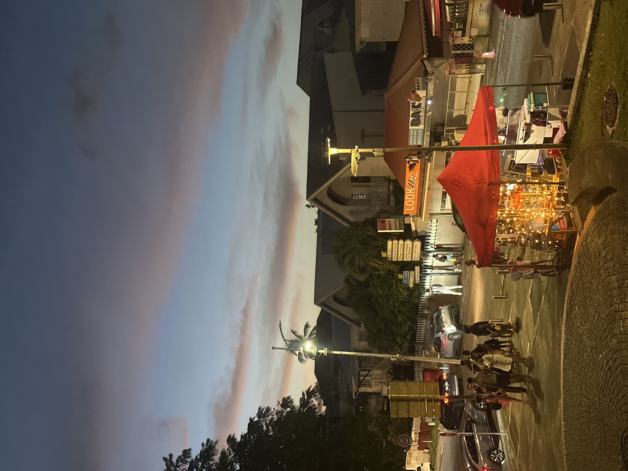 A nighttime street scene in Le Gosier, France, showcases several key elements. Under a twilight sky, a street corner is lit by streetlights. A group of people gathers, their silhouettes defined against the illuminated pavement. Cars parked along the road suggest a public space. A street vendor operates under a red awning, decorated with strings of lights. Buildings line the street, storefronts visible with their signs, including one for "Look Hair." Street signs in French direct traffic. The scene combines pedestrian activity and urban elements in the evening atmosphere of Le Gosier.