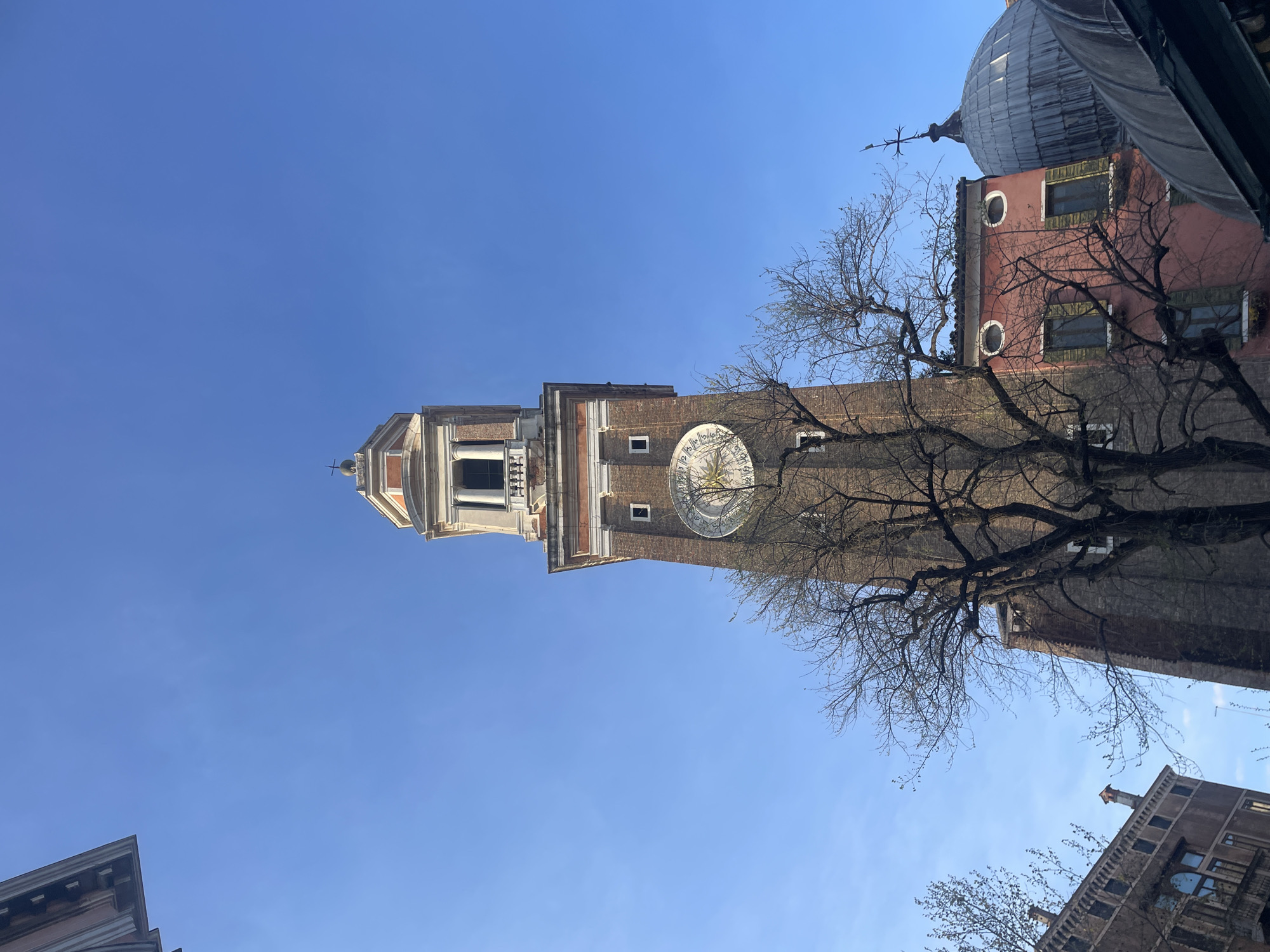 The image depicts the leaning campanile (bell tower) of Santo Stefano in Venice, Italy, partially obscured by bare tree branches. The tower, constructed of brick, features a clock face and a white-trimmed spire. To the right of the campanile, a red building with a dome-like roof is visible. The sky above is a clear, bright blue. Other buildings are seen at the bottom edges of the frame.