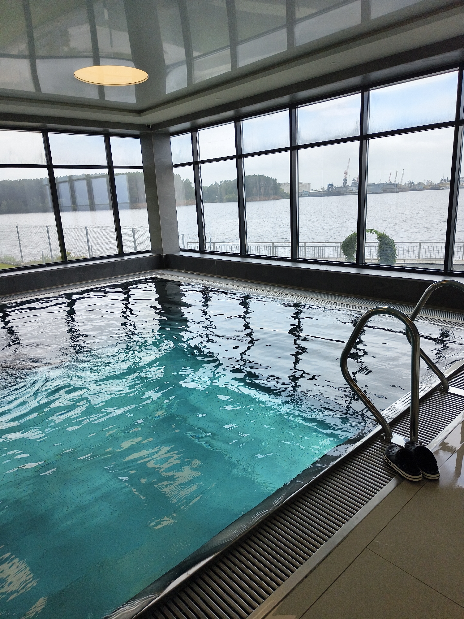 This is a medium shot of an indoor swimming pool with a view of a body of water and industrial cranes through large windows. The pool is a bright turquoise color, with visible ripples on the surface reflecting the light from the ceiling. A set of stainless steel pool ladders with two steps is visible on the right side of the pool, with a pair of black flip-flops placed next to them on the tiled floor. The pool is bordered by a dark grey edge and a silver drainage grate. The ceiling features a recessed rectangular light fixture. The large windows offer a panoramic view of a wide body of water, bordered by trees on one side and an industrial port with cranes and docked ships on the other. The sky is overcast.