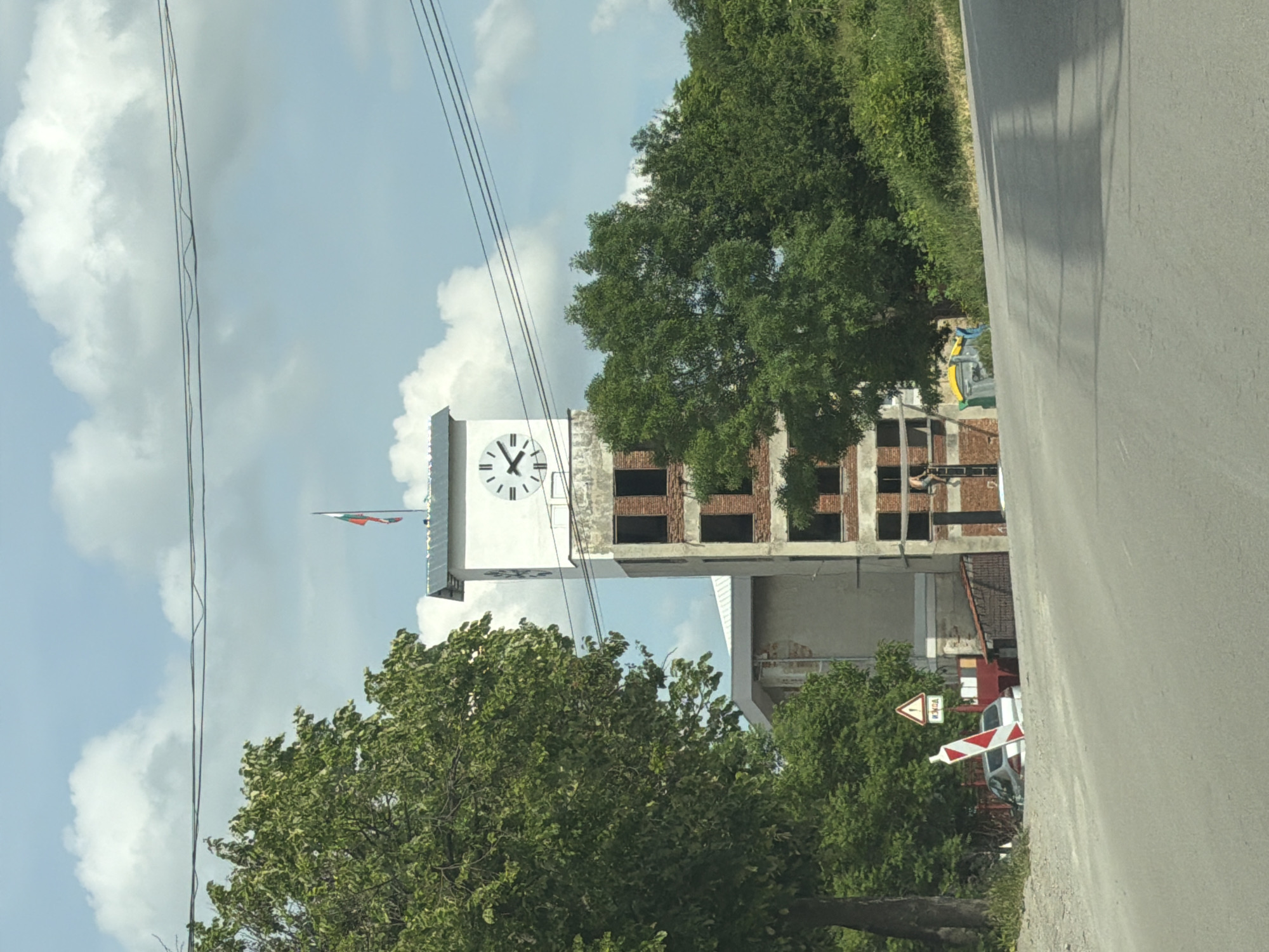 The image displays a scene featuring a prominent clock tower, adjacent buildings, and roadside elements under a bright, partly cloudy sky. The photograph appears to have been taken in a landscape orientation but is presented rotated 90 degrees counter-clockwise.
The central subject is a tall, white clock tower with a distinct black clock face, displaying Roman numerals. The clock's hands indicate approximately 11:30. A small flag, featuring the horizontal stripes of red, white, and green, is visible near the top of the tower, identifying it as the flag of Bulgaria, consistent with the location context of Shumen. Attached to or immediately beside the clock tower is a multi-story building that seems to be either under construction or undergoing significant renovation, characterized by exposed brickwork, concrete framework, and empty window openings.
In the foreground, a paved road stretches along the right side of the frame, showing shadows that suggest bright daylight, likely mid-morning or early afternoon. The road is bordered by a green verge and numerous lush trees. Several power lines crisscross the upper portion of the sky.
To the lower right, partially obscured by foliage, are roadside warning signs for a railway crossing. One sign is a triangular white sign with a red border, containing the number '1' and Cyrillic text below it that reads "ЖП ПРЕЛЕЗ," which translates to "Railway Crossing" (ЖП is an abbreviation for "Железопътен," meaning "railway"). The number '1' typically indicates a single track. Adjacent to this is a red and white striped barrier or chevron sign, commonly used to mark the approach to a railway crossing.