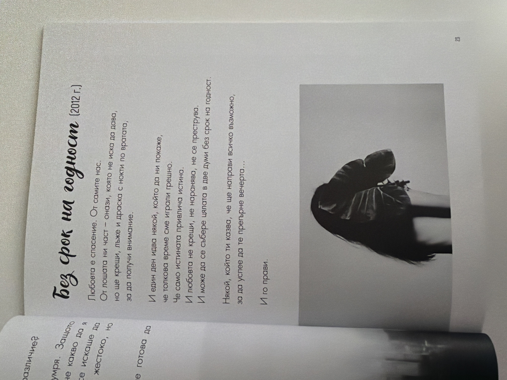 The image shows an open book with text on the left page and text and a black and white photograph on the right page. The left page contains partially visible text in Bulgarian. The right page has a title in a decorative font that reads "Без срок на годност (2012 г.)" which translates to "No expiration date (2012)". Below the title are several lines of Bulgarian text, followed by a black and white photograph of a woman from the chest up, facing away from the camera. She has long dark hair and is wearing a dark garment, possibly a jacket with a collar. The photograph is framed by what appears to be large, dark green leaves. The bottom right corner of the page shows the number "23". The image does not contain any specific elements that would indicate its location in Pazardzhik, Bulgaria.