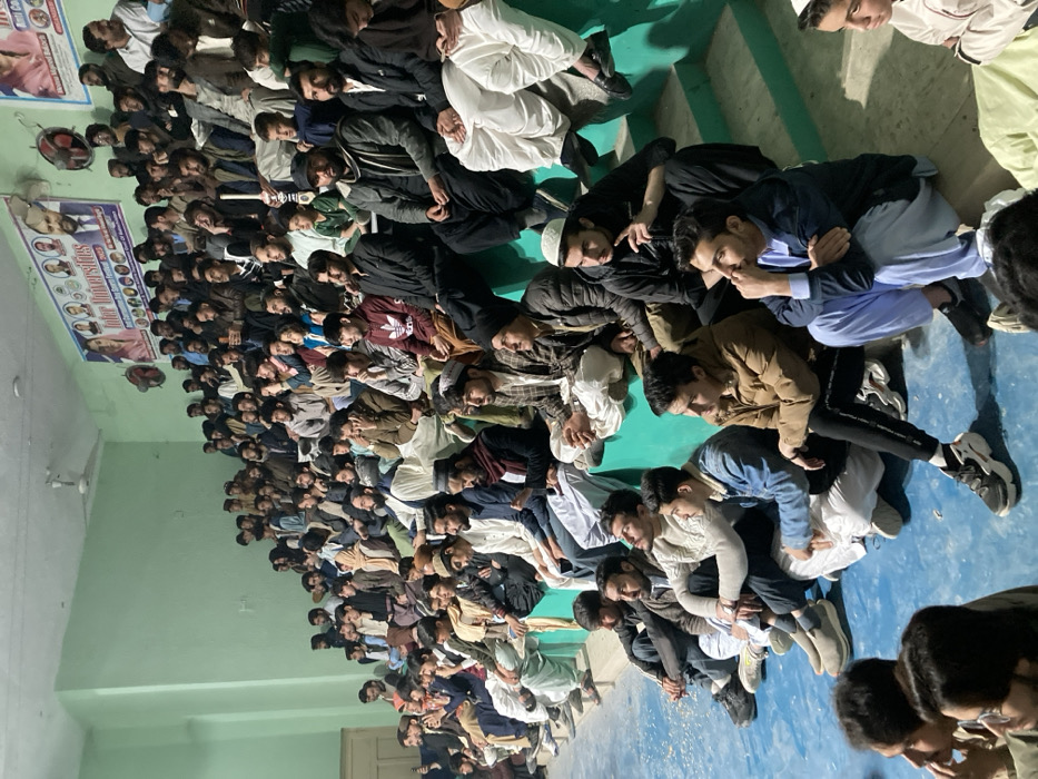 The image depicts an indoor scene within a large room, identified as being located in Quetta City Tehsil, Pakistan. The room features light green walls and a blue floor, which appears to be a court surface, with scattered white debris on it. Tiered seating, constructed from light green steps, occupies the right and background portions of the frame. A white ceiling fan is visible overhead.
On the far wall, two large banners are displayed. The banner on the left, largely visible, features images of men and women alongside the text "Inter Universities All Commercial Sports Festival 2023." The banner on the right, partially visible, displays images of men. Two wall-mounted fans are positioned on the back wall near the banners. A light-colored wooden door is partially visible on the left wall.
A large gathering of approximately 100 to 150 men and boys, ranging from youth to middle-aged adults, occupies the tiered seating and also sits directly on the blue floor in the foreground. Their attire primarily consists of traditional Pakistani clothing such as Shalwar Kameez in various colors (white, beige, blue, black) worn with jackets, sweaters, or hoodies. Some individuals wear skullcaps (topis), while others are in Western-style jeans, t-shirts, and sneakers.
The individuals are primarily in a seated or crouching position, facing an unseen central area, indicating their role as spectators. Their expressions vary from attentive observation to more relaxed or contemplative states. No active group interaction or conversation is prominently depicted; most individuals appear to be independently observing. One young man in the upper middle section holds a white stick-like object. One individual in the lower right foreground is looking at a mobile phone.