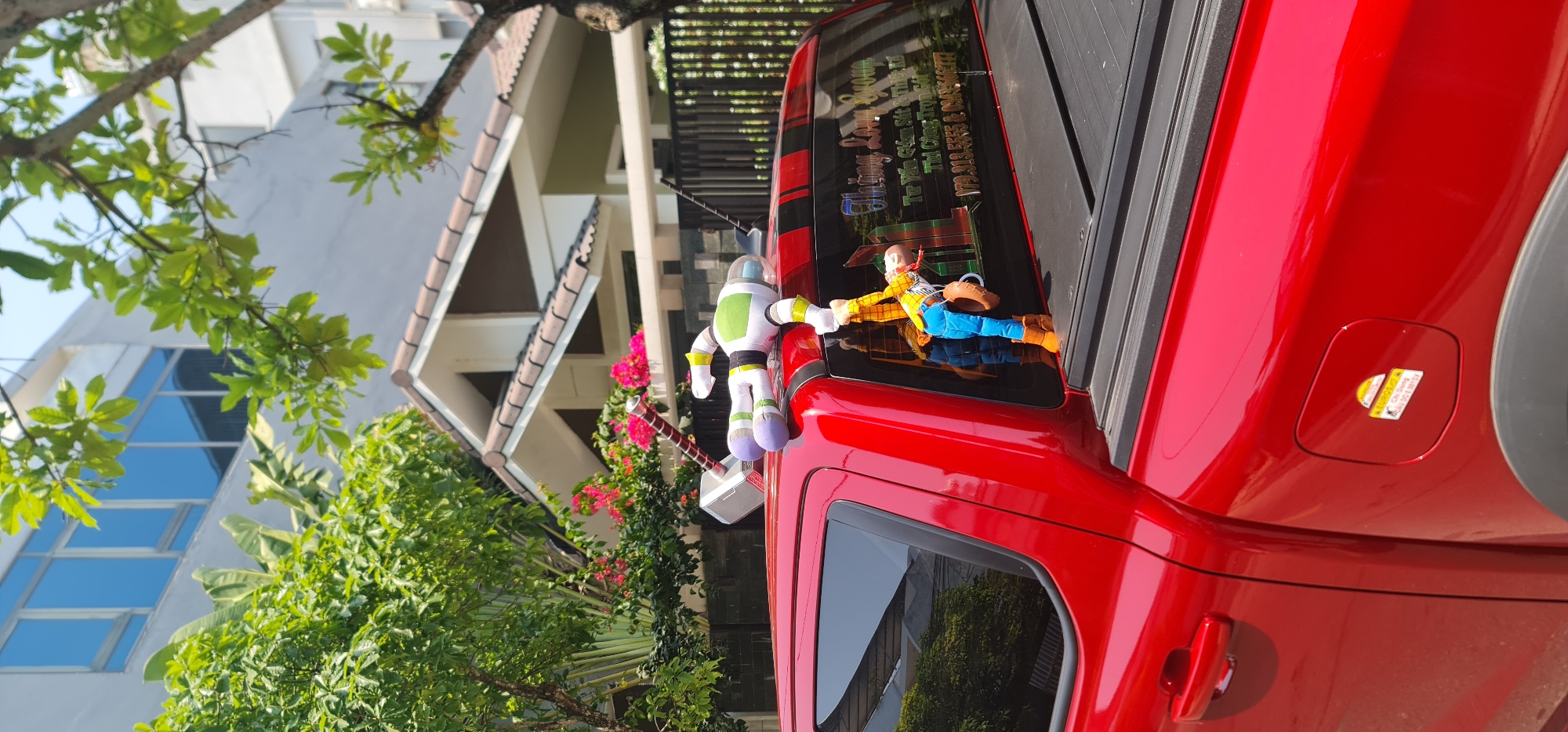 The image depicts a red car parked outdoors, likely in a residential area in Đà Nẵng, Vietnam. The car is adorned with two "Toy Story" plush toys, Buzz Lightyear and Woody, positioned on the roof above the window. Buzz Lightyear is holding a toy hammer, seemingly Mjolnir. The car's rear window has stickers with Vietnamese writing. The background includes greenery, a building with a light-colored facade, and a glimpse of a blue-tinted window. The scene appears to be captured on a bright, sunny day.