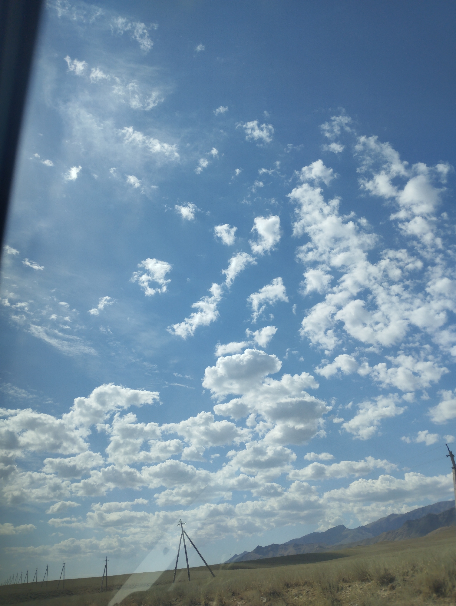 The image captures a vast expanse of blue sky filled with scattered white cumulus clouds. In the foreground, a dry, grassy plain stretches towards a range of low, undulating mountains in the distance. A line of utility poles with crossbars runs along the horizon, suggesting an open, possibly rural or undeveloped area. The lighting indicates it is daytime, likely with the sun positioned high in the sky. The scene is devoid of any human figures or visible structures apart from the utility poles. The overall impression is one of a wide-open landscape under a dynamic sky. The provided context mentions "Temirkavuk, Uzbekistan," which would imply this scenery is representative of that geographical location.