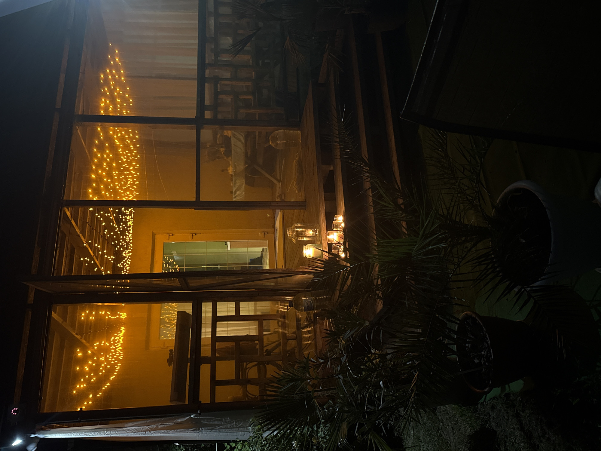 The image depicts the exterior of a screened-in porch or gazebo at night in Serhiivka, Ukraine. The structure is illuminated by strings of warm-toned fairy lights draped across the ceiling. Inside, a table is set, suggesting a dining or gathering space. Several glass lanterns with lit candles are placed on the wooden steps leading up to the porch, casting a soft glow. Potted plants, including palm-like foliage, are situated in the foreground. The overall atmosphere is cozy and inviting, despite the darkness. The presence of the fairy lights and candles creates a festive or romantic ambiance.