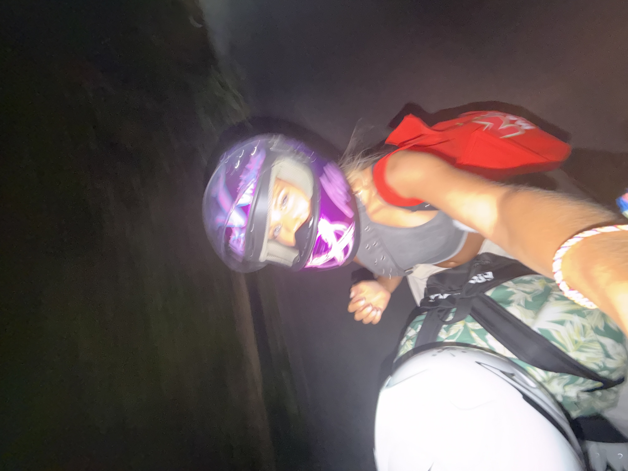 A woman wearing a purple and silver motorcycle helmet with a reflective visor is riding on the back of a motorcycle at night. She has blonde hair and is wearing a grey top. A red bag is slung over her right shoulder. In front of her is another motorcycle helmet, white in color, and a green and white patterned bag. The background is dark, indicating a nighttime scene, possibly on a road in North Kuta, Indonesia. The camera angle suggests the photo was taken from the perspective of the motorcycle driver.
