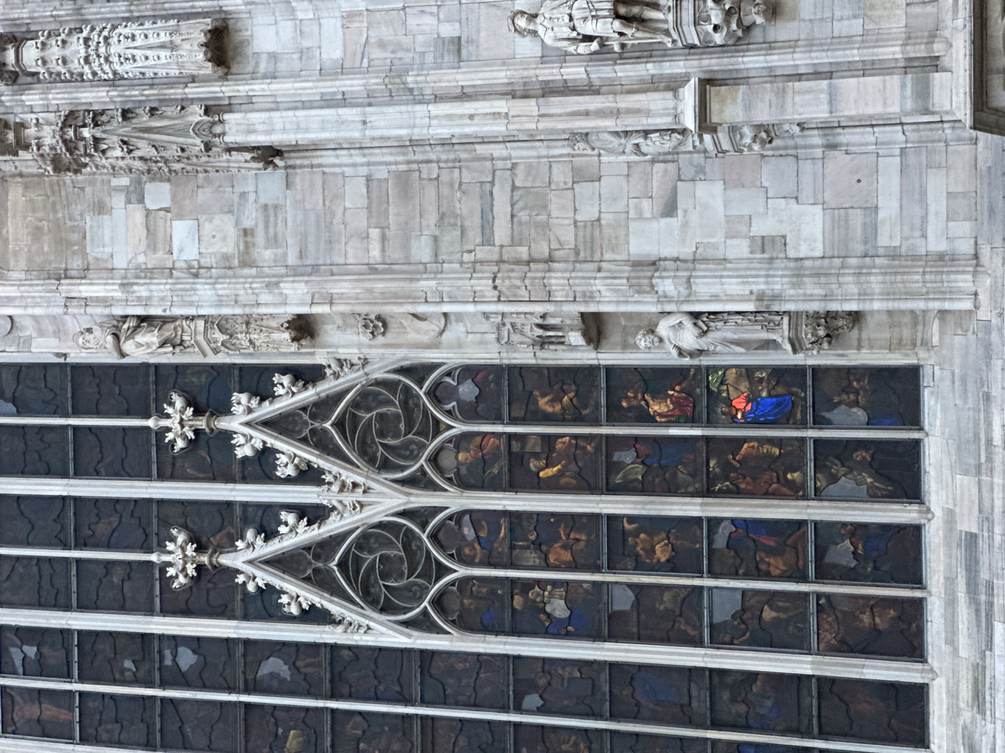 The image shows a close-up view of a section of the Duomo di Milano, a Gothic cathedral located in Milan, Italy. The scene features a tall, narrow stained-glass window with multiple panels depicting figures in colorful scenes. The window is framed by ornate white stonework, including pointed arches and decorative elements. To the right of the window, the stone facade of the cathedral is visible, characterized by textured, light-colored stone blocks and architectural details such as columns, niches, and statues. One statue, a male figure in classical attire, stands prominently in a niche on the right side of the image. The overall impression is one of intricate architectural detail and historical significance.