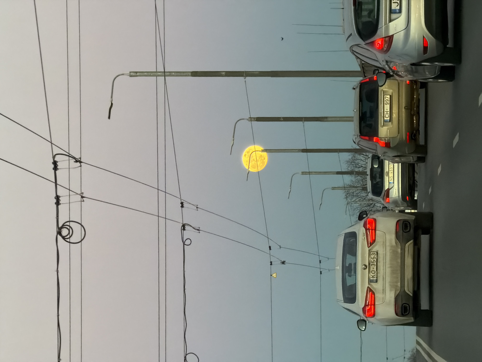 The image shows a street scene in Riga, Latvia. A line of cars is visible on the right side of the frame, including a white BMW with license plate "KO-3553," a gold-colored car with license plate "CH-997," and a silver car. Overhead power lines and streetlights are prominent in the left and center portions of the frame. A bright yellow moon is visible between the streetlights.