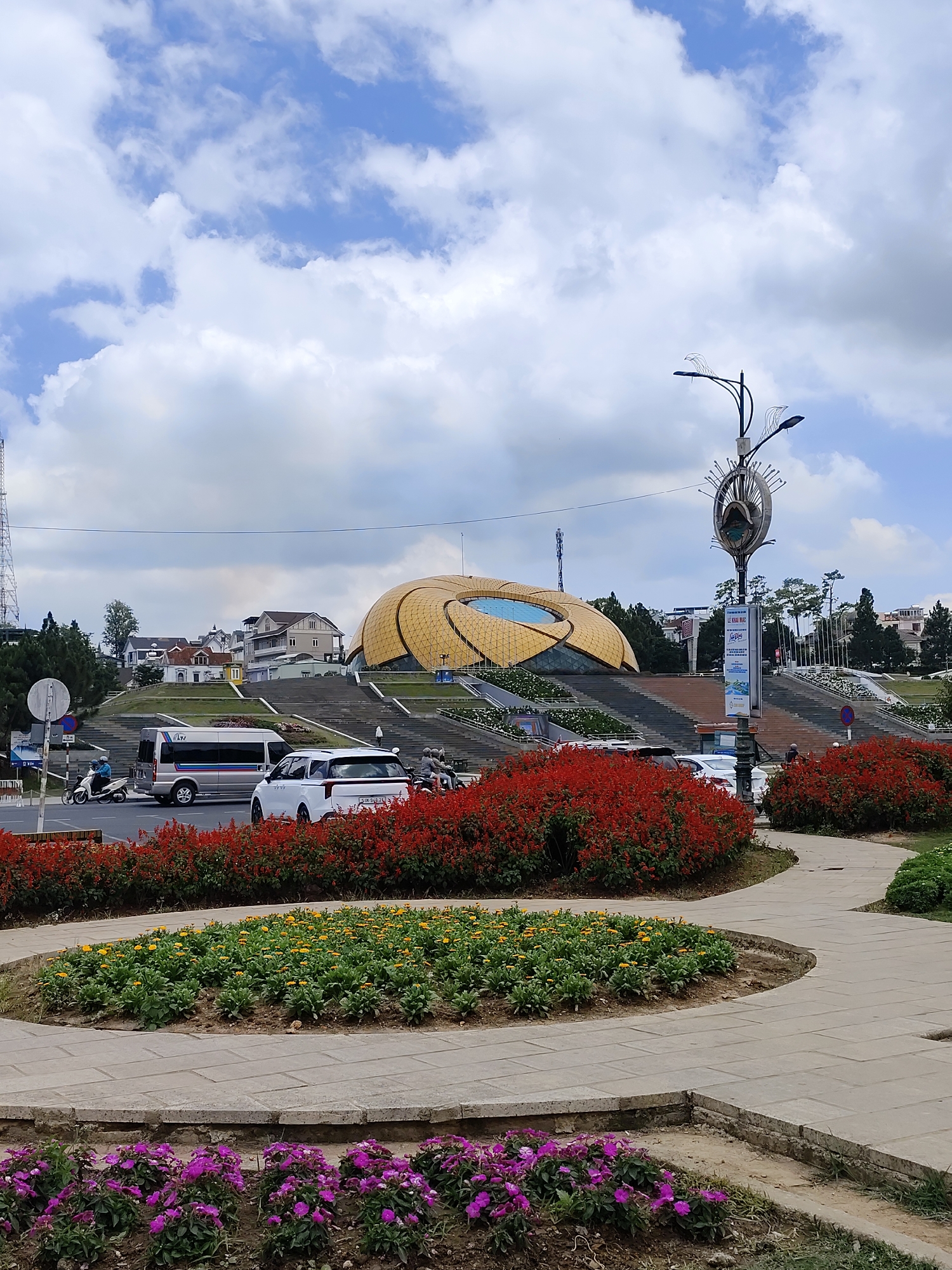 The image shows a sunny day in Da Lat, Vietnam. In the background is the Golden Bee building, a prominent landmark in the city. Below the Golden Bee, there are sets of steps leading to the city center. In the foreground, there are colorful flower beds with red, yellow, and purple flowers. A gray stone pathway winds through the flower beds. Several vehicles are visible in the midground, including a white van and a white car. A streetlight with decorative elements stands on the right. The sky is partly cloudy.