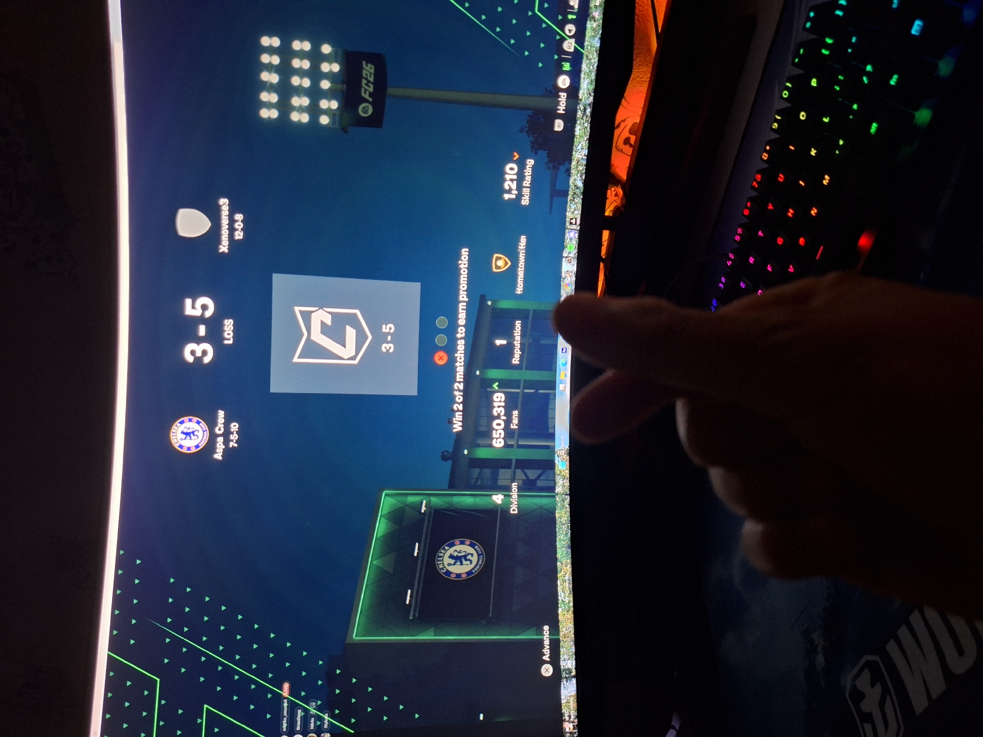 The image shows a close-up of a person's hands forming a finger heart gesture in front of a computer monitor displaying a video game interface, specifically FC 26. The game screen shows a match score of 3-5 with "LOSS" indicated. Details on the screen include team names "Aspa Crew" and "Xenoverse3", along with their respective scores and win-loss records. The Chelsea Football Club logo is also visible. The environment is dark, with the computer monitor and a keyboard with RGB lighting being the primary light sources. The person is positioned such that their hands and a portion of their lap are visible in the foreground, obscuring the lower part of the monitor and the keyboard. There is no information in the image to suggest any specific location in the city of Varna, Bulgaria.