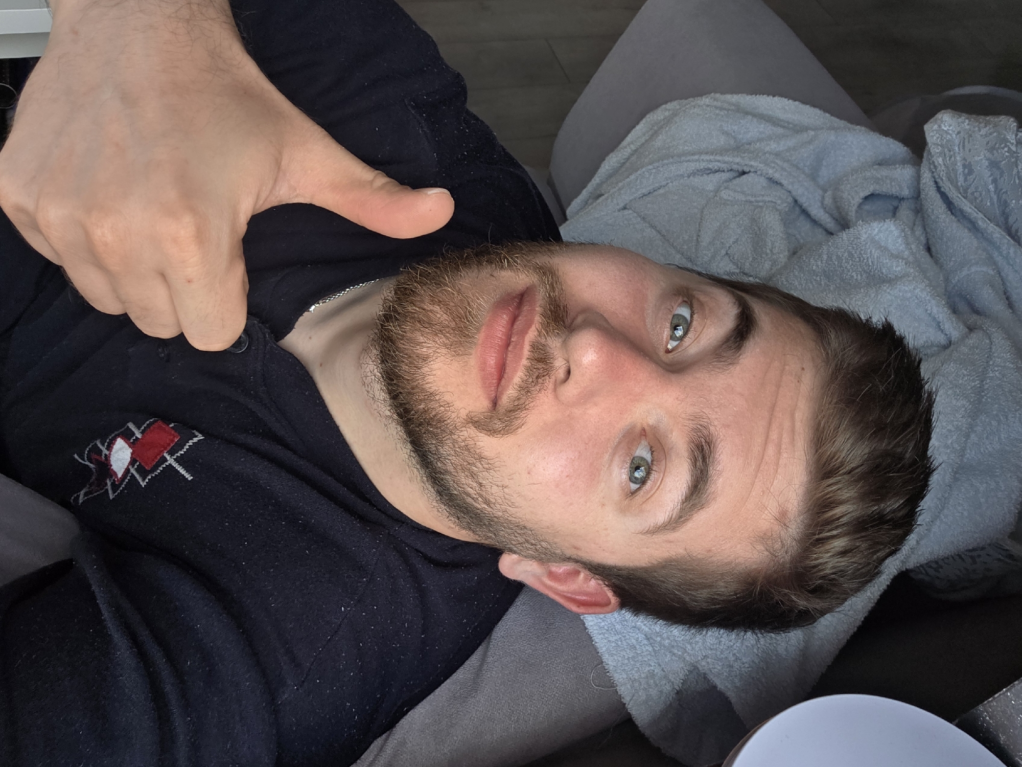 A man is lying on a gray sofa or cushion, with a light blue towel or blanket draped behind his head. He is wearing a dark blue shirt with a red and white logo near his shoulder. He has a beard and light blue eyes. His right hand is raised, giving a thumbs-up gesture. The background shows what appears to be a light-colored floor. The location is not definitively discernible as Yagodovo, Bulgaria, based on the image alone.