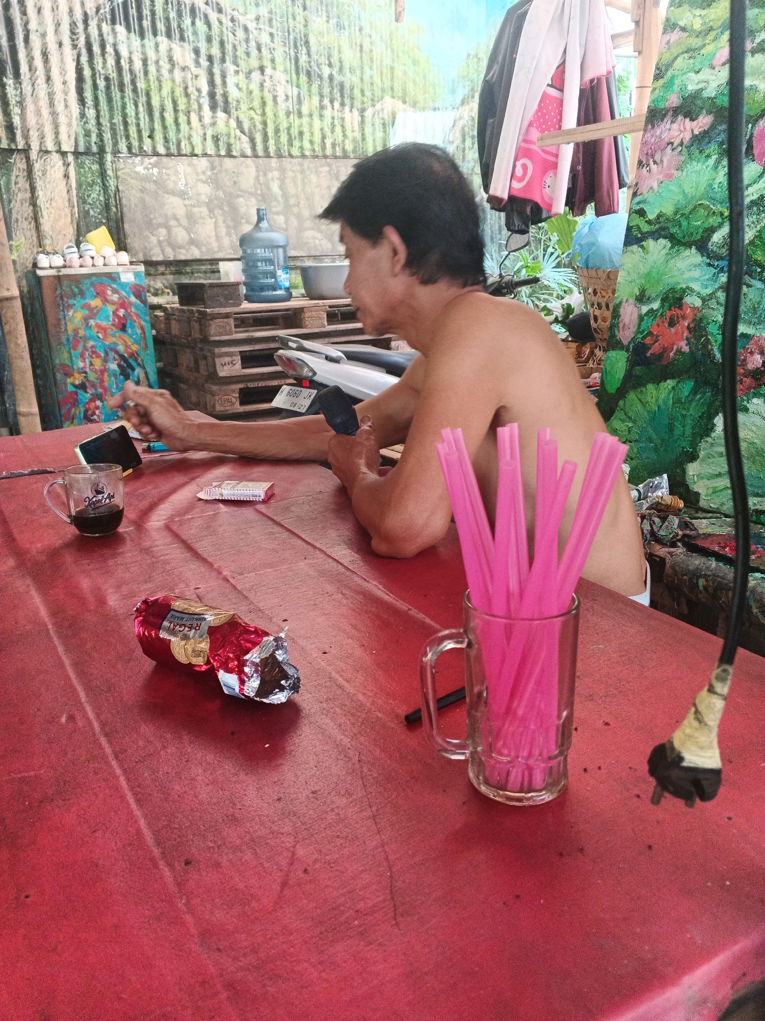 The image captures a man sitting at a red, outdoor table. He appears to be in Ungaran, Indonesia, as indicated by the context provided. The man, shirtless and with short black hair, is looking down at his hands. One hand is holding a dark-colored object, possibly a microphone or a small electronic device. In front of him on the table are several items: a glass mug with a dark liquid, a pack of cigarettes, and a partially opened candy wrapper. To his right, a clear glass mug filled with pink drinking straws stands prominently. Behind the man, the environment is somewhat cluttered, with paintings, a large water jug, and what appears to be motorcycle parts visible. The overall impression is of a casual, perhaps informal, outdoor setting.