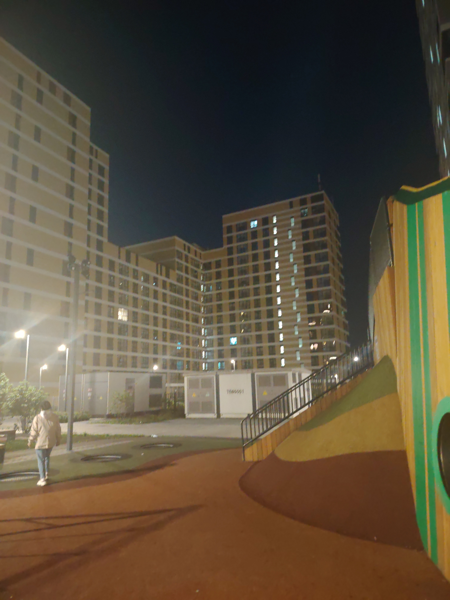 The image shows a nighttime scene with a person walking away from the camera on a brown, rubberized surface. In the background, there are several large, modern apartment buildings illuminated by artificial lights. The buildings have a consistent pattern of windows and balconies. To the right, a colorful, curved structure with green and yellow stripes and a circular opening is visible. The overall environment appears to be an urban residential area, possibly a park or a common space within a housing complex, at night. The specific location in Yekaterinburg, Russia, is not identifiable from the visual information alone.