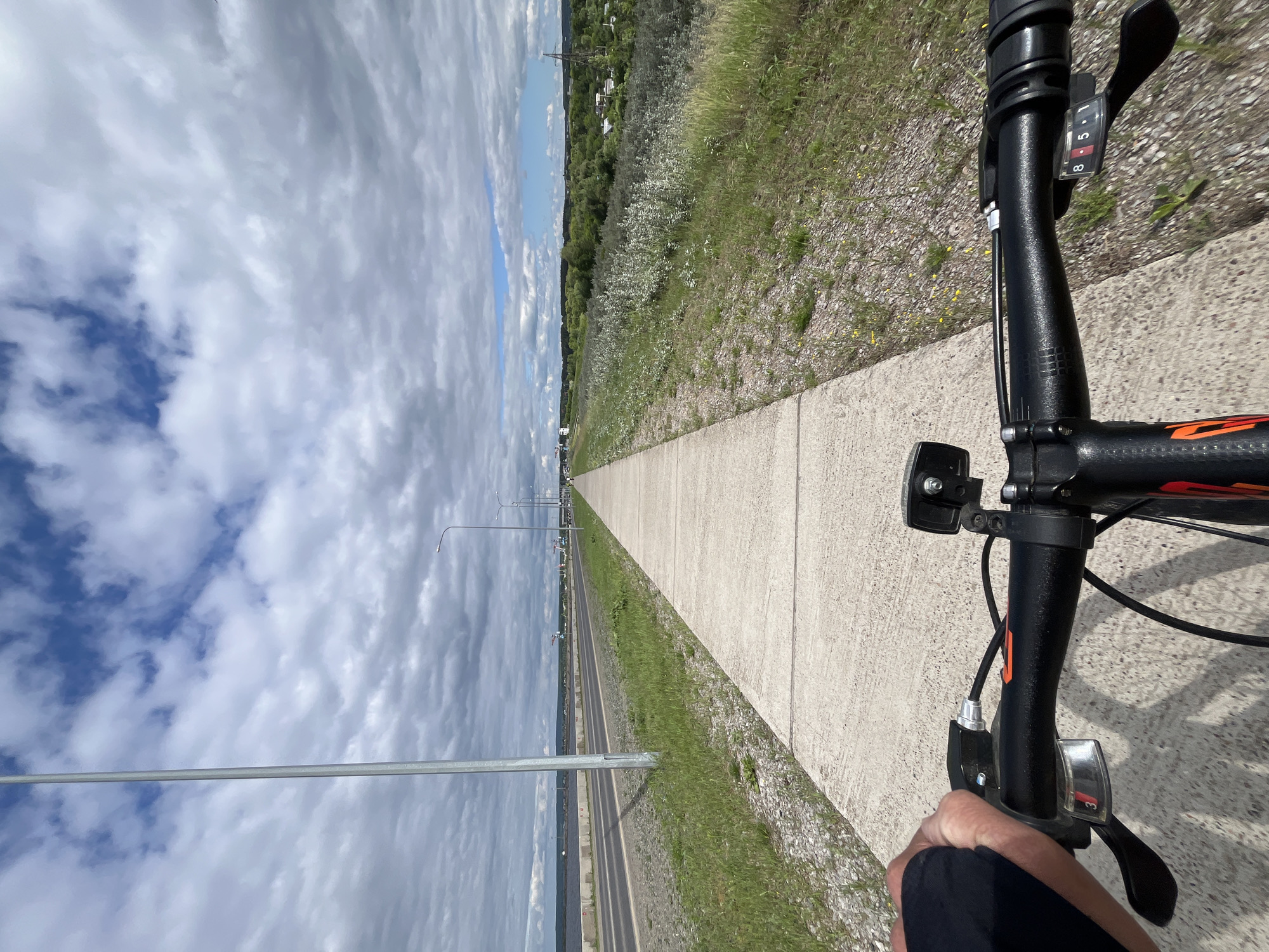 The image shows a first-person perspective of a person riding a bicycle on a paved path. The path runs alongside a road and a body of water, likely the Kama River, with a cityscape visible in the distance. The sky is cloudy with patches of blue. The bicycle handlebars are visible in the foreground, with a hand gripping the left handlebar. The scene appears to be in the city of Chaikovsky, Russia, possibly along the embankment of the Kama River near the port, given the industrial elements and the wide expanse of water. The environment suggests a partly urban and partly natural setting on a cloudy day.