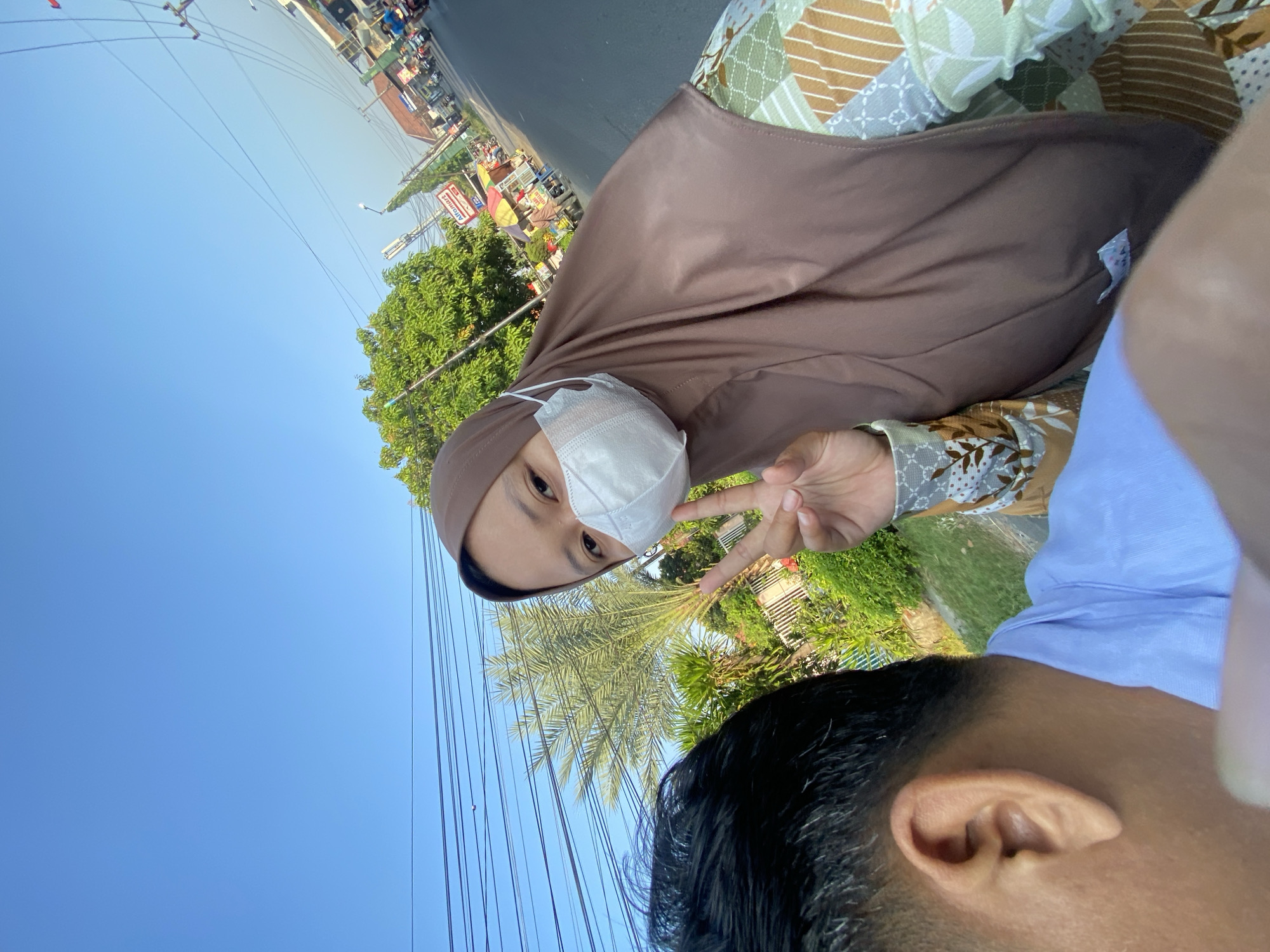 A selfie taken from the perspective of a person riding on the back of a motorcycle. The rider in the foreground has black hair and is wearing a light blue shirt. To their right, a person wearing a brown hijab and a white face mask is looking towards the camera and making a peace sign with their right hand. They are wearing a green and brown patterned blouse. Behind them, a palm tree and lush green foliage are visible, along with a street lined with buildings and shops. In the distance, the sky is clear and blue, with overhead power lines crisscrossing the frame. The scene appears to be in a residential or commercial area on a sunny day. The image shows a glimpse of daily life on a street in Balonggabus, Indonesia.