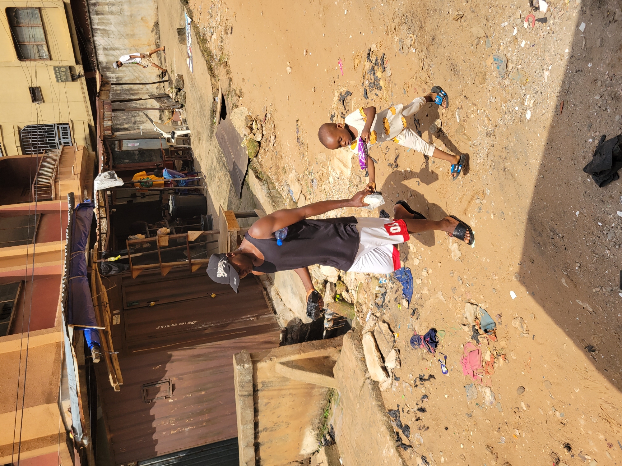 In the city of Onitsha, Nigeria, a man wearing a black tank top, white shorts with red trim, a black baseball cap, and black sandals walks on a dirt path littered with debris. He holds a plastic bag in his right hand and a white object, possibly a bottle of water, in his left. Beside him, a young child in a light-colored outfit with cloud patterns and blue sandals walks, holding a purple object. The background features a building with a corrugated metal roof and an awning. The ground is predominantly dirt and sand, with scattered refuse including plastic bags, clothing, and rocks. Sunlight casts shadows on the ground.
