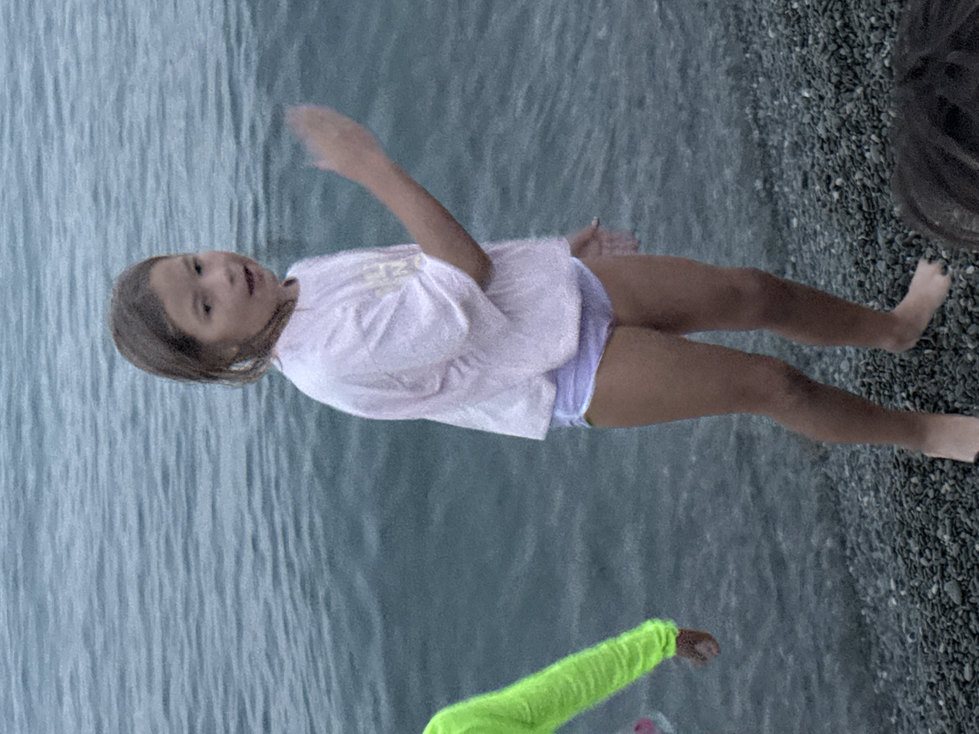 The image depicts two young girls on a pebble beach next to a body of water. The girl in the foreground is wearing a light pink shirt and purple shorts. Her body is angled towards the water, with her right arm extended upwards and her left arm slightly bent. She has brown hair and appears to be looking towards the water. She is standing on a pebble beach, with the water to her left.
Behind her, another child is visible from the waist down, wearing a bright green long-sleeved shirt. Their left hand is extended towards the viewer. This child is also on the pebble beach.
The water has a textured appearance, suggesting ripples or waves. The pebble beach is composed of dark-colored stones. The overall lighting suggests it might be an overcast day or late afternoon, as there are no strong shadows. The setting appears to be a natural outdoor environment, likely a beach or shoreline. No text is visible in the image.