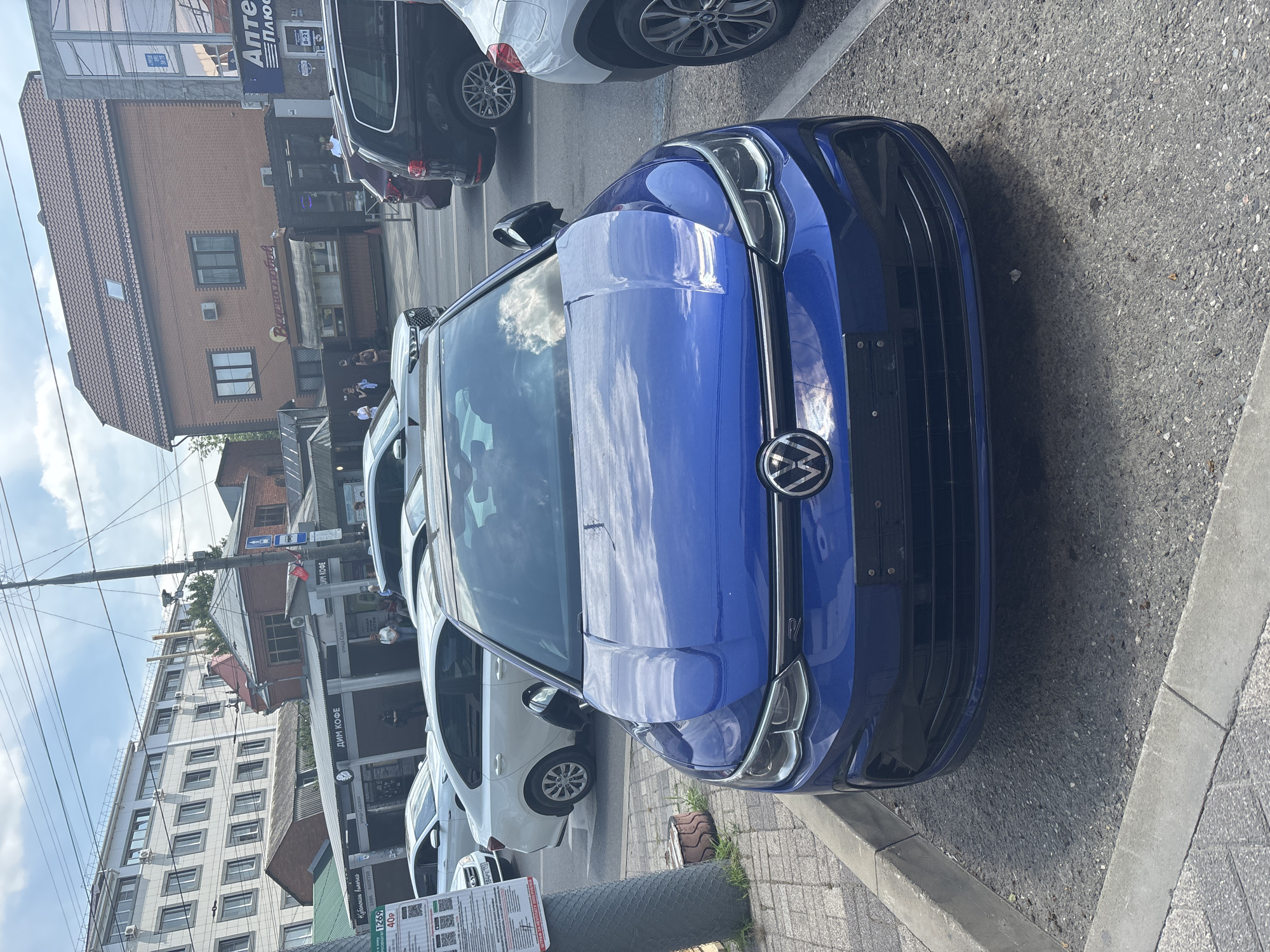 The image shows a blue Volkswagen Golf parked on an asphalt lot next to a sidewalk in Krasnodar, Russia. In the background, a brick building with a red roof houses shops and businesses, including "Аптека Плюс" (Apteka Plus, meaning Pharmacy Plus) and "Вкуснотище" (Vkusnotishche, meaning Deliciousness). Other cars are visible in the background, including a white sedan, a dark-colored SUV, and a white SUV. The sky is partly cloudy. The parking area is enclosed by concrete curbs, and a sign is attached to a pole on the left side of the image, displaying information in Russian.