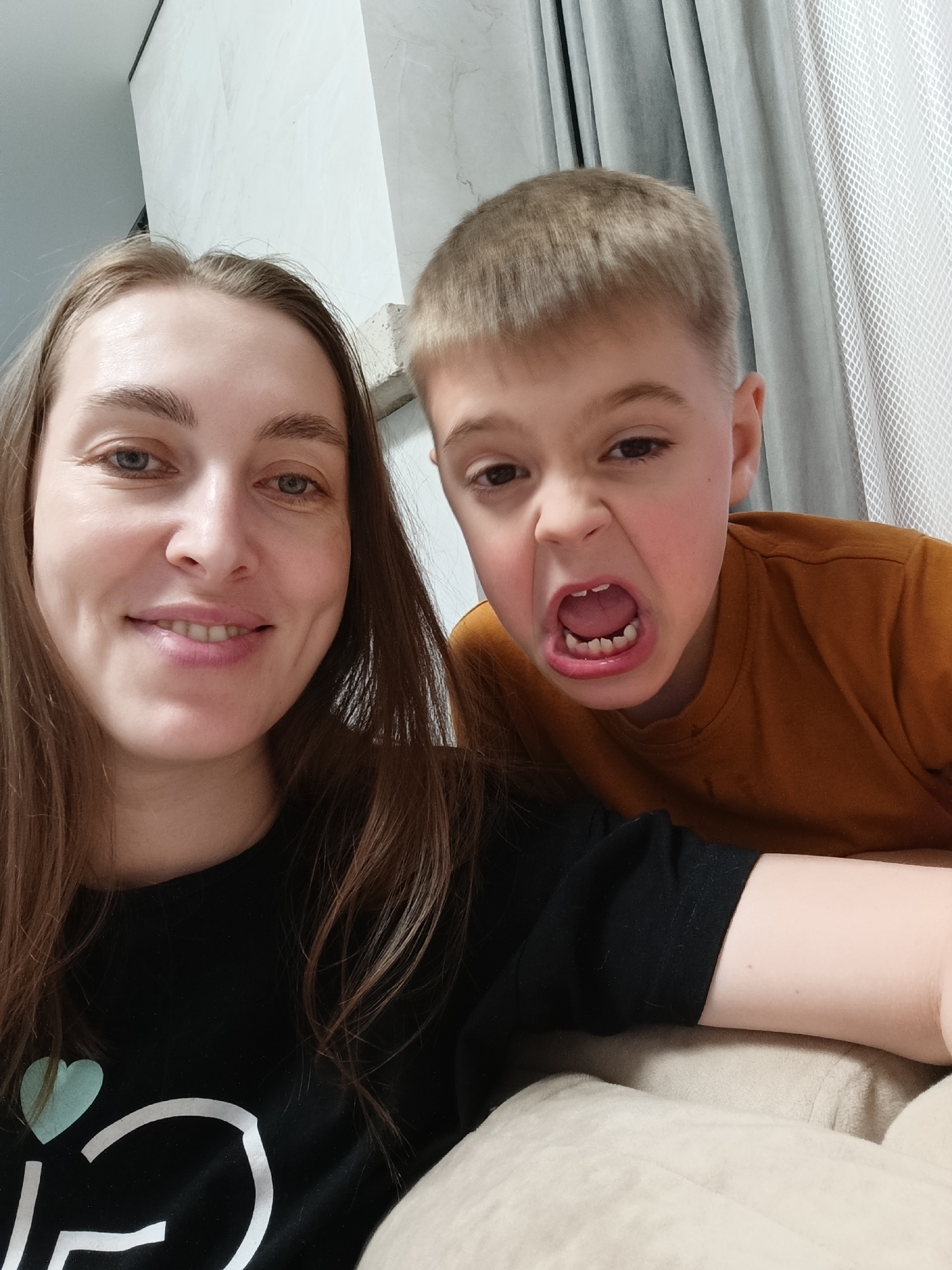 A selfie photo taken indoors shows a woman and a young boy. The woman, with long brown hair, is smiling at the camera. She is wearing a black t-shirt with a white design and a turquoise heart. The boy is behind her, making a face with his mouth open, showing his teeth. He has short blond hair and is wearing an orange t-shirt. The background includes light-colored walls and curtains. The specific location in the city of Chaikovsky, Russia, cannot be determined from the image.