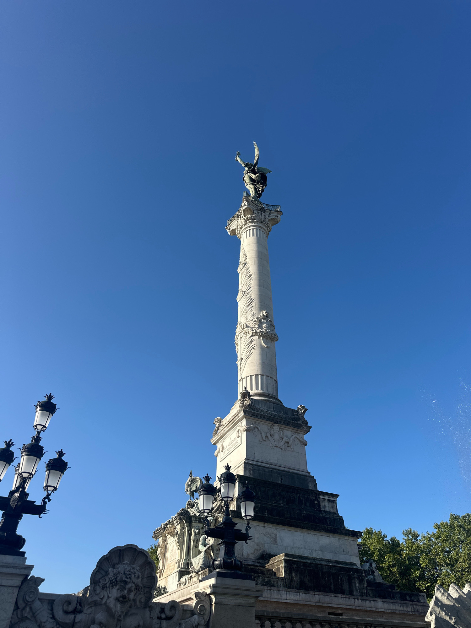 This is a low-angle, full-shot photograph of the Monument aux Girondins in Bordeaux, France. The monument is a tall, white stone column topped by a statue of the "Spirit of Democracy." The column is intricately decorated with sculptures and bas-reliefs. In the foreground to the left, a black lamppost with multiple globes is visible, and to the right, a section of trees and a fountain spray are present. The sky is a clear, bright blue. The monument is situated in a public square.