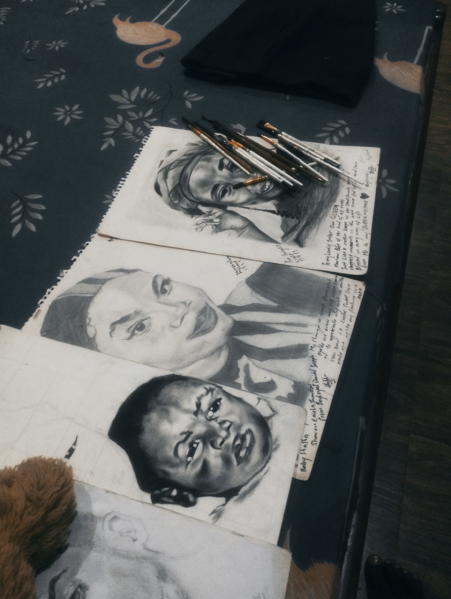 A close-up, overhead shot captures several charcoal sketches laid out on a patterned fabric surface. The fabric features a repeating pattern of stylized leaves and a single orange flamingo. Resting on top of the sketches are a collection of paintbrushes with metal ferrules and wooden handles. In the background, a dark, folded object, possibly clothing or a book, is visible.
The sketches themselves appear to be portraits. The topmost sketch shows a person wearing a headscarf. Below this, a larger sketch depicts the face of a woman with prominent eyebrows and lips. The bottom sketch features a baby's face. Faint handwritten text is visible on some of the sketches. A small portion of a brown, fuzzy object, resembling a stuffed animal, is visible in the lower left corner. The lighting is dim, casting subtle shadows.