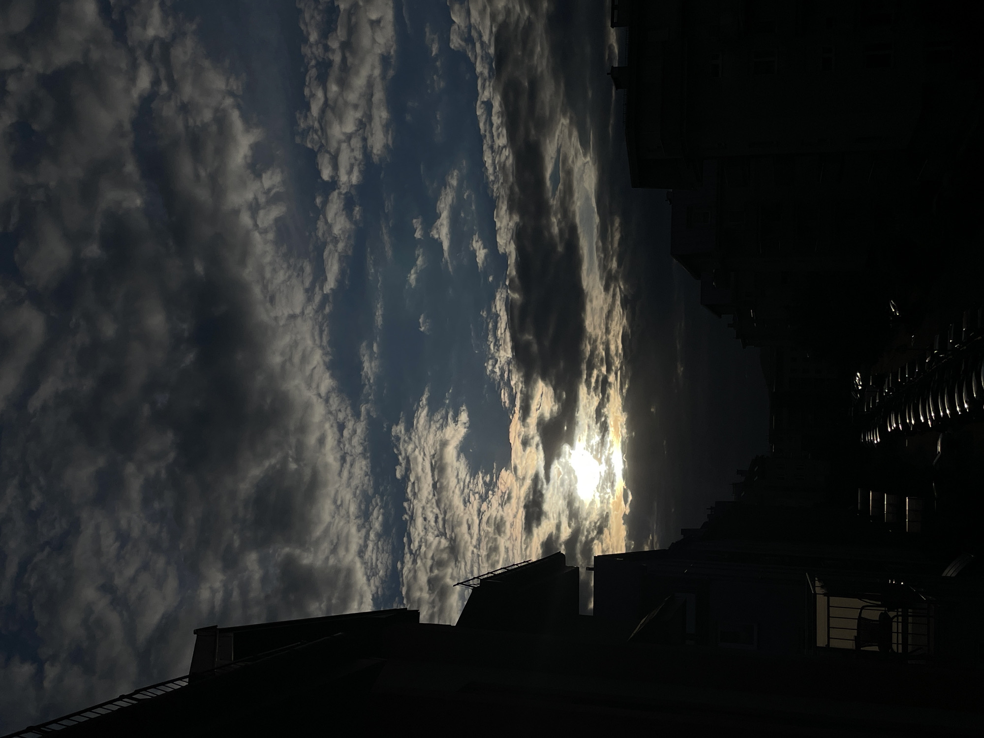 The image depicts a street scene at dusk, with the sun setting behind a building, casting long shadows. The sky is filled with dramatic, dark clouds, interspersed with patches of lighter sky. The buildings on either side of the street are silhouetted against the sky, with one building on the left featuring a balcony with a chair. In the foreground, rows of parked cars are visible, also in silhouette. The overall impression is one of a moody, atmospheric urban environment. The location is identified as Unknown, Unknown.