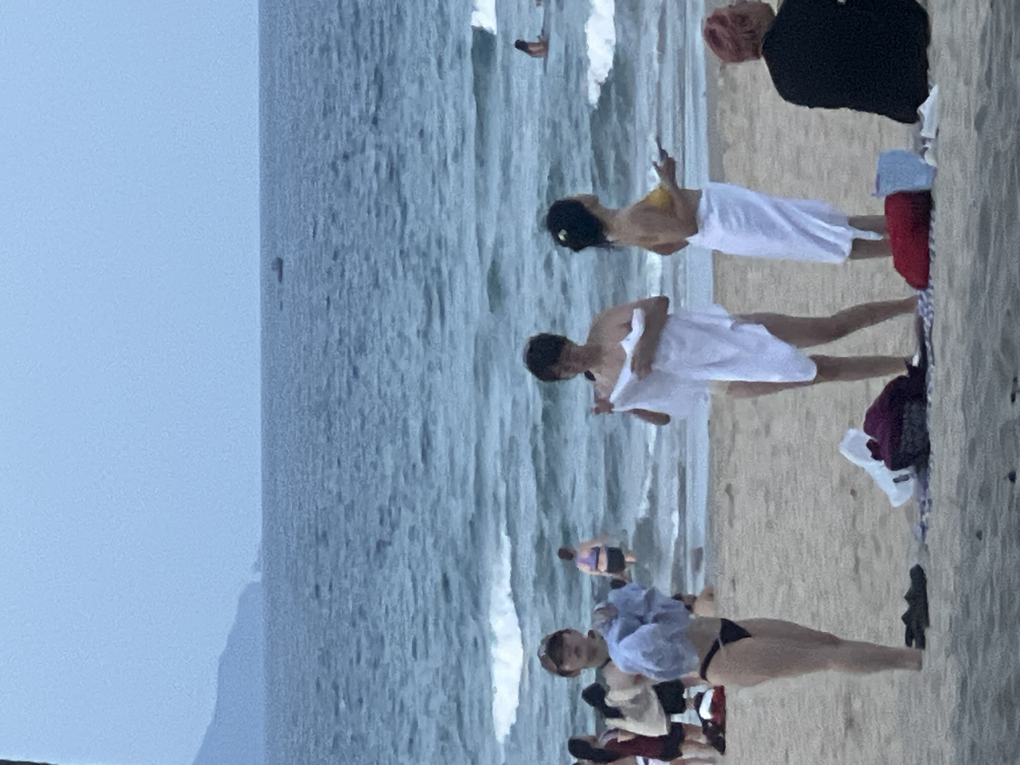 In Đà Nẵng, Vietnam, at a beach with moderate waves and a hazy sky, several individuals are present. On the sandy shore, a person wearing a black bikini and a cap is holding a light blue towel. To their right, another person wrapped in a white towel holds another white towel. Further to the right, a person in a yellow bikini top and white towel is holding a phone. In the background, several other people are in the water, and a small boat is visible on the horizon. The scene depicts a typical day at the beach with people engaged in various activities.