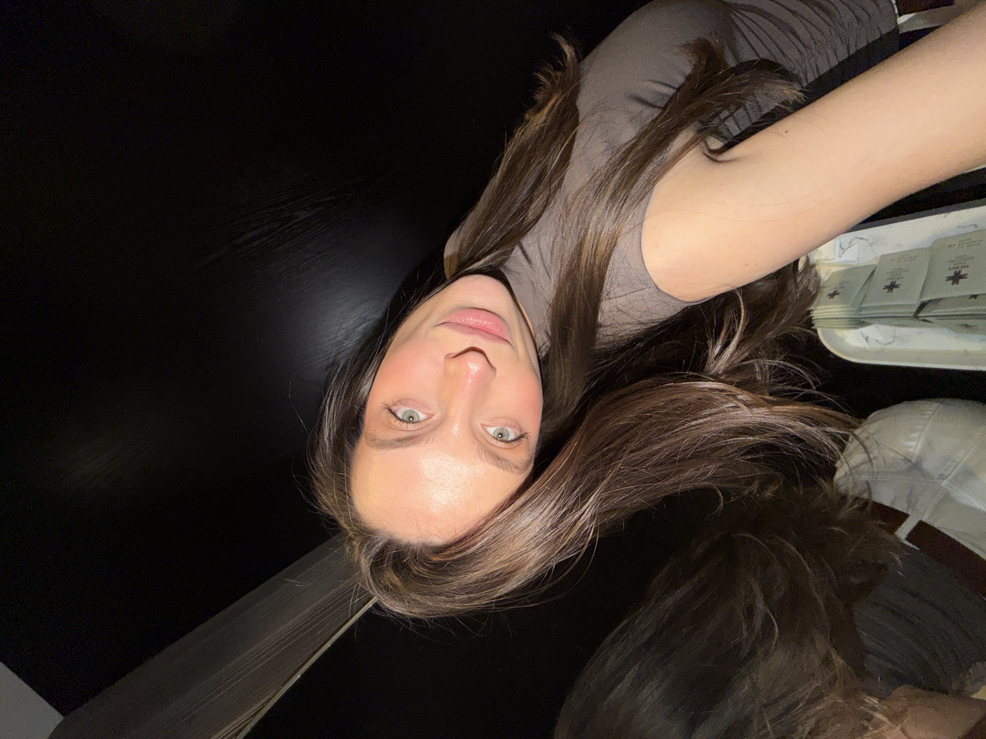A selfie image from a low angle of a young woman with long brown hair and fair skin. She is wearing a brown t-shirt. To the left, the image features a dark, black surface, possibly a table. To the right, there is a tray with what appears to be small packaged items with a cross symbol on them. In the background, a white hat is visible.
The image provides insufficient information to confidently ascertain the location of the subject.