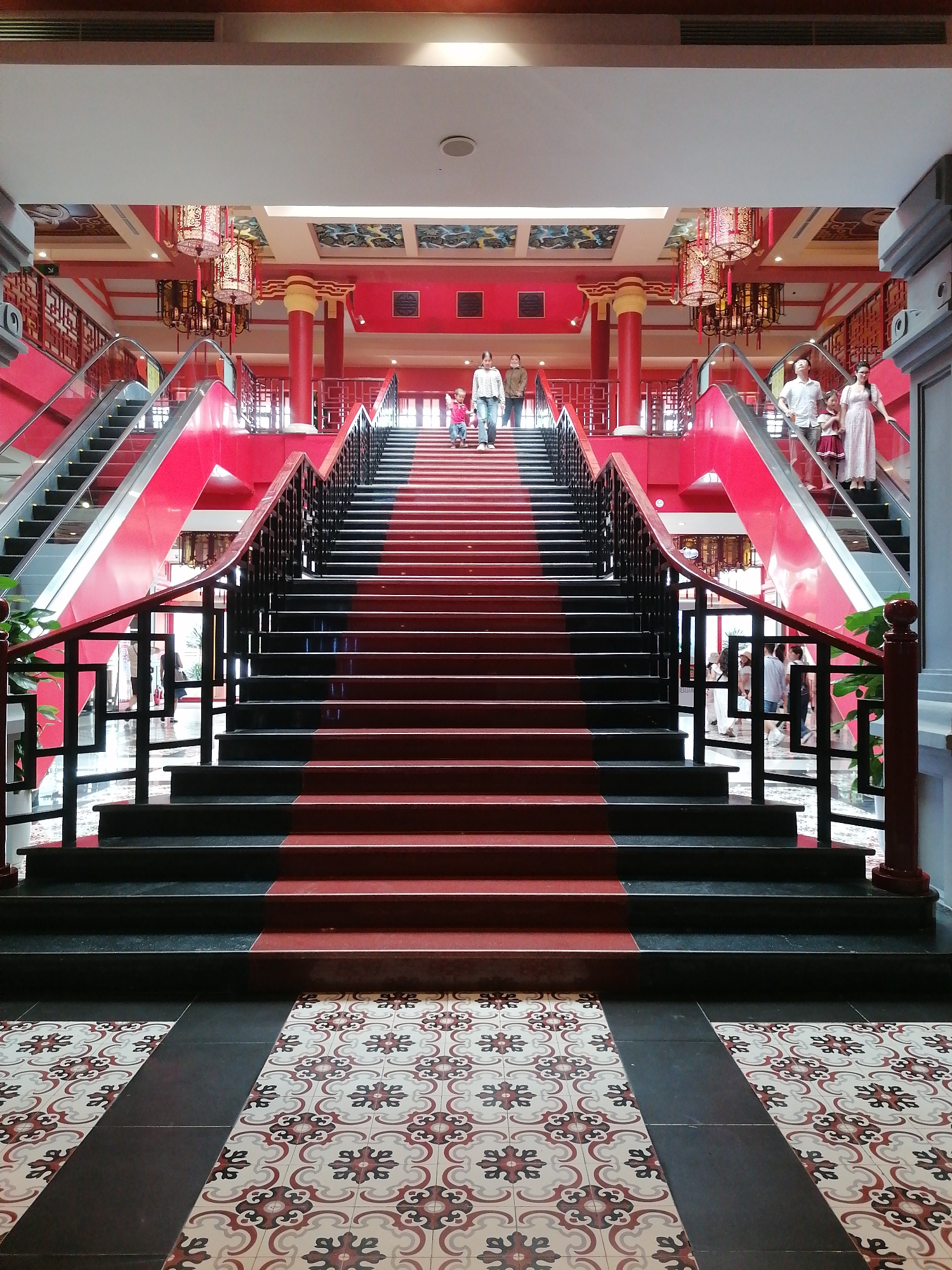 The image depicts a grand staircase and escalators in a building with Chinese architectural influences, likely in Bai Chay Ward, Vietnam. The staircase, covered in a red runner, ascends to an upper level, flanked by escalators on either side. People are seen on both the stairs and escalators. Red is the predominant color, with black accents on the stairs and railings. The floor is tiled with a patterned design in red, white, and black. The building features red pillars and decorative lanterns.