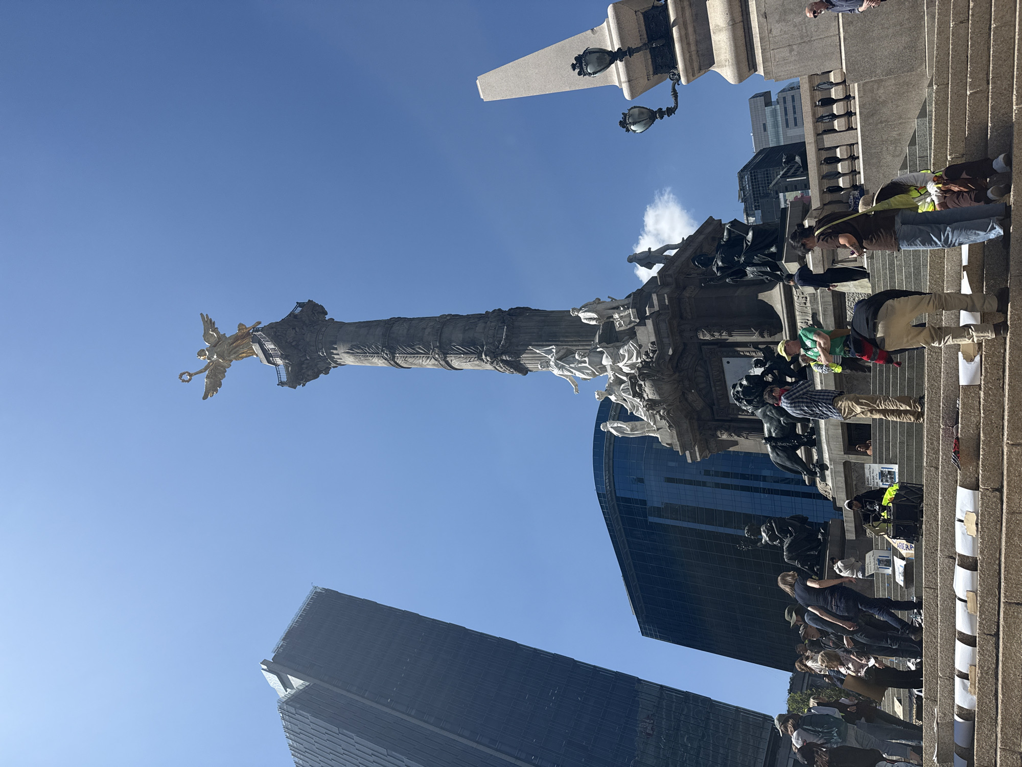 The image depicts the Angel of Independence monument in Mexico City, Mexico, from a low-angle perspective. The monument, topped with a golden angel statue, dominates the frame against a clear blue sky. Modern skyscrapers rise in the background, suggesting the monument's location in a bustling urban area. At the base of the monument, a crowd of people is present, some standing on the steps. Signs are propped up on the stairs, indicating a gathering or protest of some kind. The scene is captured in daylight, with sunlight illuminating the monument and casting shadows.
