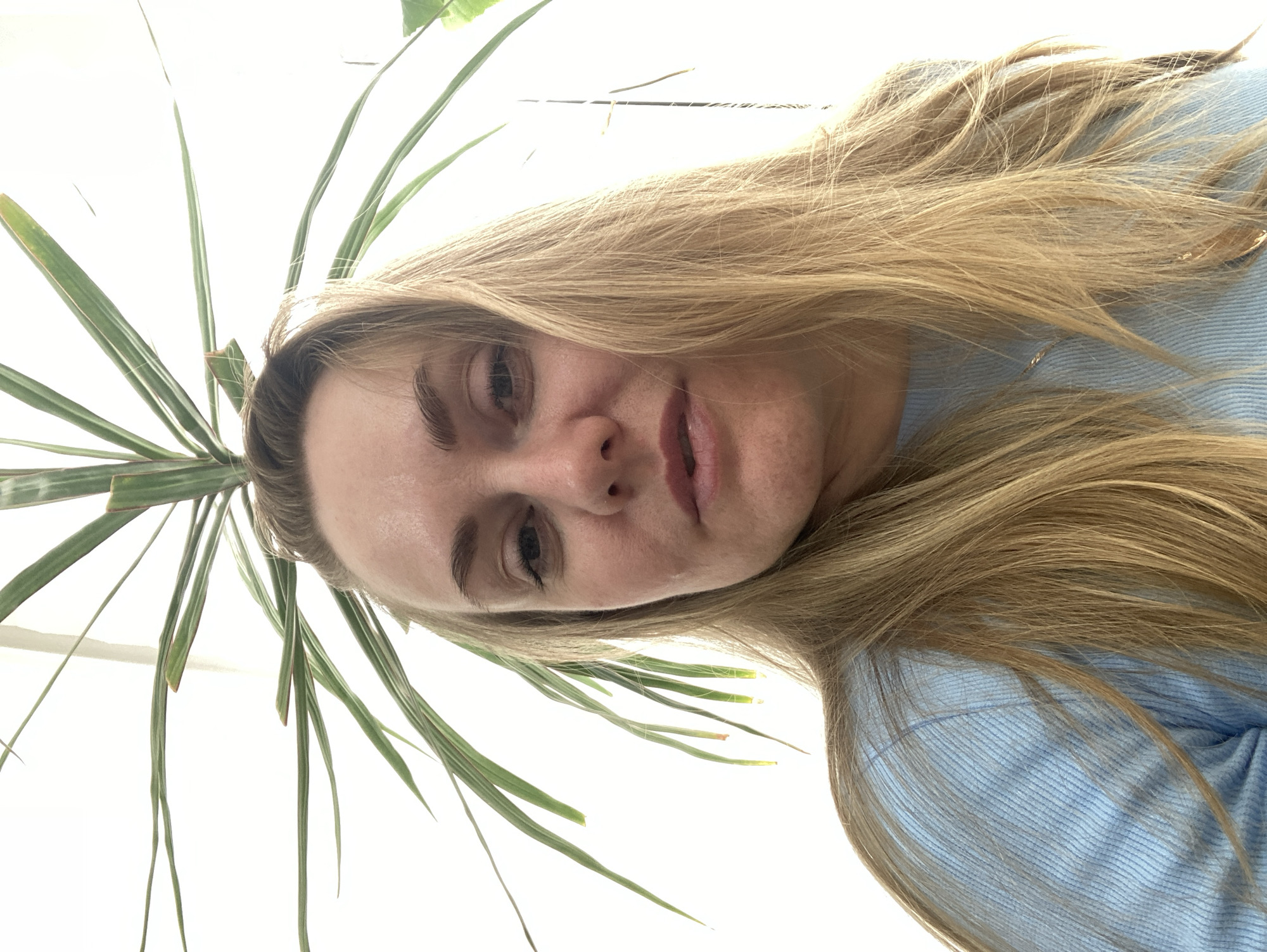 A rotated, low-angle portrait captures a young adult woman with long, blonde hair against a bright white background, framed by the vivid green leaves of an indoor plant. The woman's face is partially visible, tilted to her right, as she looks directly at the camera with a neutral, calm expression. Her lips are slightly parted, and her eyes are open. She has fair skin and appears to be wearing a light blue, ribbed long-sleeved top or sweater, visible on the right side of the frame.
Several long, sword-shaped green leaves, consistent with an indoor plant such as a dracaena or similar palm species, surround her head, particularly prominent in the upper and left sections of the image. The background is a stark, bright white, suggesting an overexposed window, a ceiling, or a plain wall, indicating an indoor setting.
The photograph is taken from a very low vantage point, looking upwards at the subject, and is presented rotated approximately 90 degrees counter-clockwise, creating an intimate and unconventional perspective. The bright background implies good daylight. No text is visible in the image. This scene is set indoors, possibly in Chaikovsky, Russia, as per context.