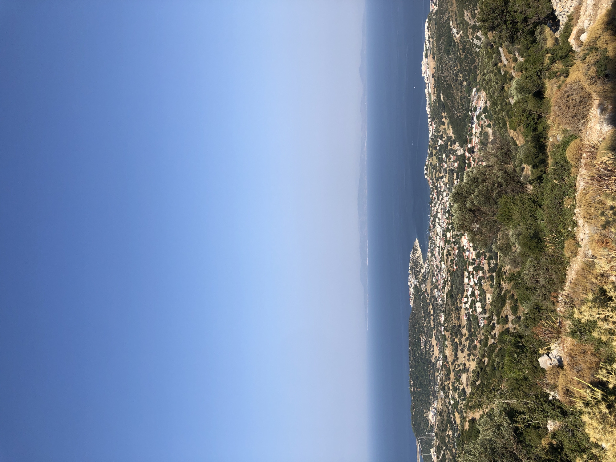 This is a high-angle, full shot of a coastal town nestled among rolling hills overlooking a vast expanse of the Aegean Sea, under a clear blue sky. The foreground features dry, scrubby vegetation and rocky terrain, with some olive trees scattered throughout. The middle ground reveals a densely populated town with white-roofed buildings, interspersed with green trees and winding roads. Further in the distance, the deep blue sea stretches to the horizon, where faint, hazy islands can be seen. The scene appears to be a panoramic view of Karaburun, Turkey, likely taken from a high vantage point. There are no people or animals visible in the image.