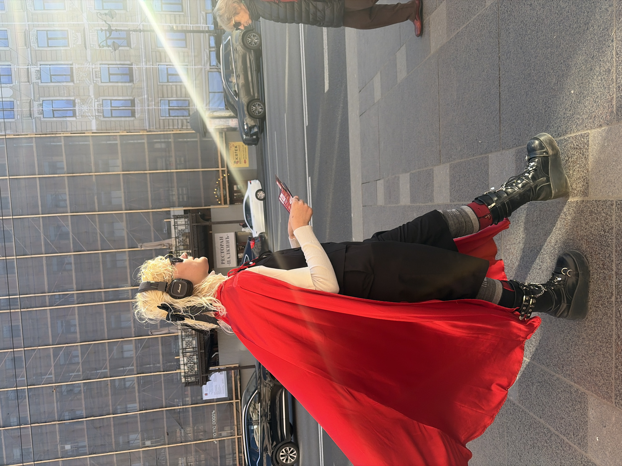 A female individual with blonde hair, wearing headphones and a red cape, is standing on a sidewalk in Saint Petersburg, Russia, holding a smartphone. She is wearing black boots, black pants, and a white long-sleeved shirt. A building with scaffolding is visible in the background, and cars are parked on the street. Another person is visible in the upper right corner of the frame.