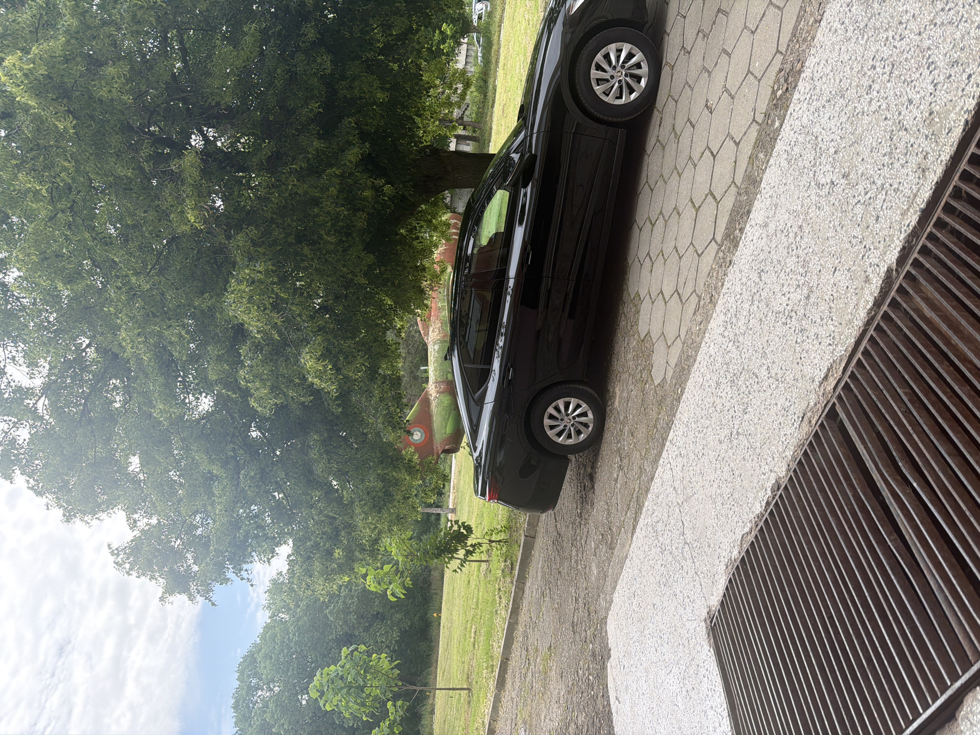 A black car is parked on a paved area adjacent to a grassy field with trees. In the background, there is an old red and green airplane on display. A metal grate is visible in the foreground. The location is Dolna Mitropolia, Bulgaria.