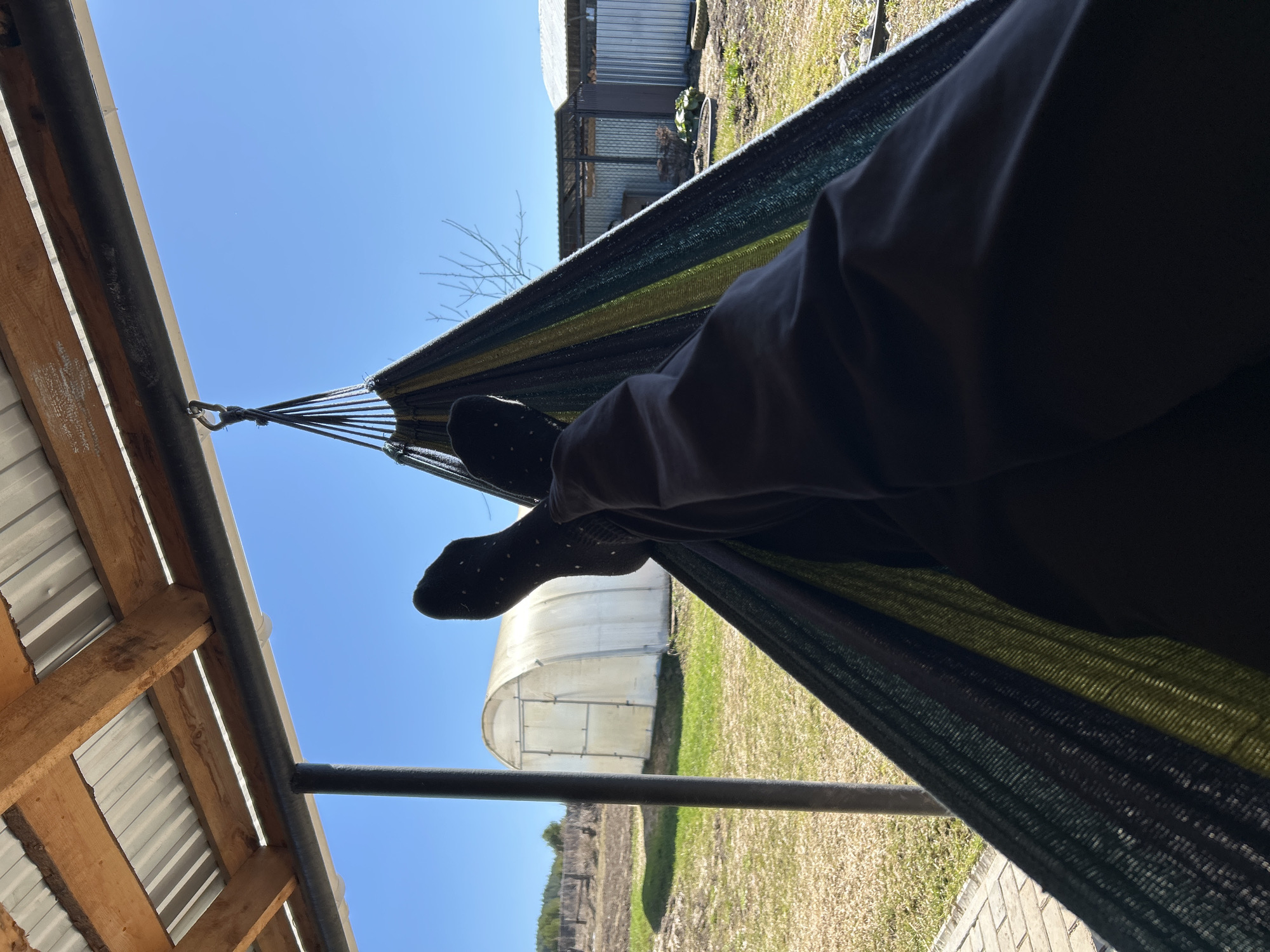 The image shows a person lying in a hammock. The hammock is suspended between a metal support structure and a building. The person is wearing dark pants and black socks with white polka dots. The view from the hammock includes a blue sky, a greenhouse, a metal shed, and a grassy area. The location is Уральское, Russia.