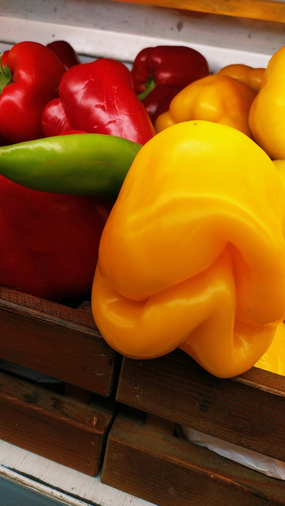 In Horishni Plavni, Ukraine, this close-up shot captures a vibrant display of bell peppers. The peppers are arranged in what appears to be wooden crates, suggesting a market or produce stand. The primary subjects are red, yellow, and green bell peppers. A large, prominently featured yellow bell pepper dominates the foreground, its smooth, curved surface reflecting the light. Behind it, a red bell pepper is visible, partially obscured by the yellow one, and further back, more red and yellow peppers are visible. A sliver of a bright green pepper is also present, adding another color to the composition. The wooden crates are weathered and appear to be stacked. The overall lighting suggests an indoor setting or a shaded outdoor area.