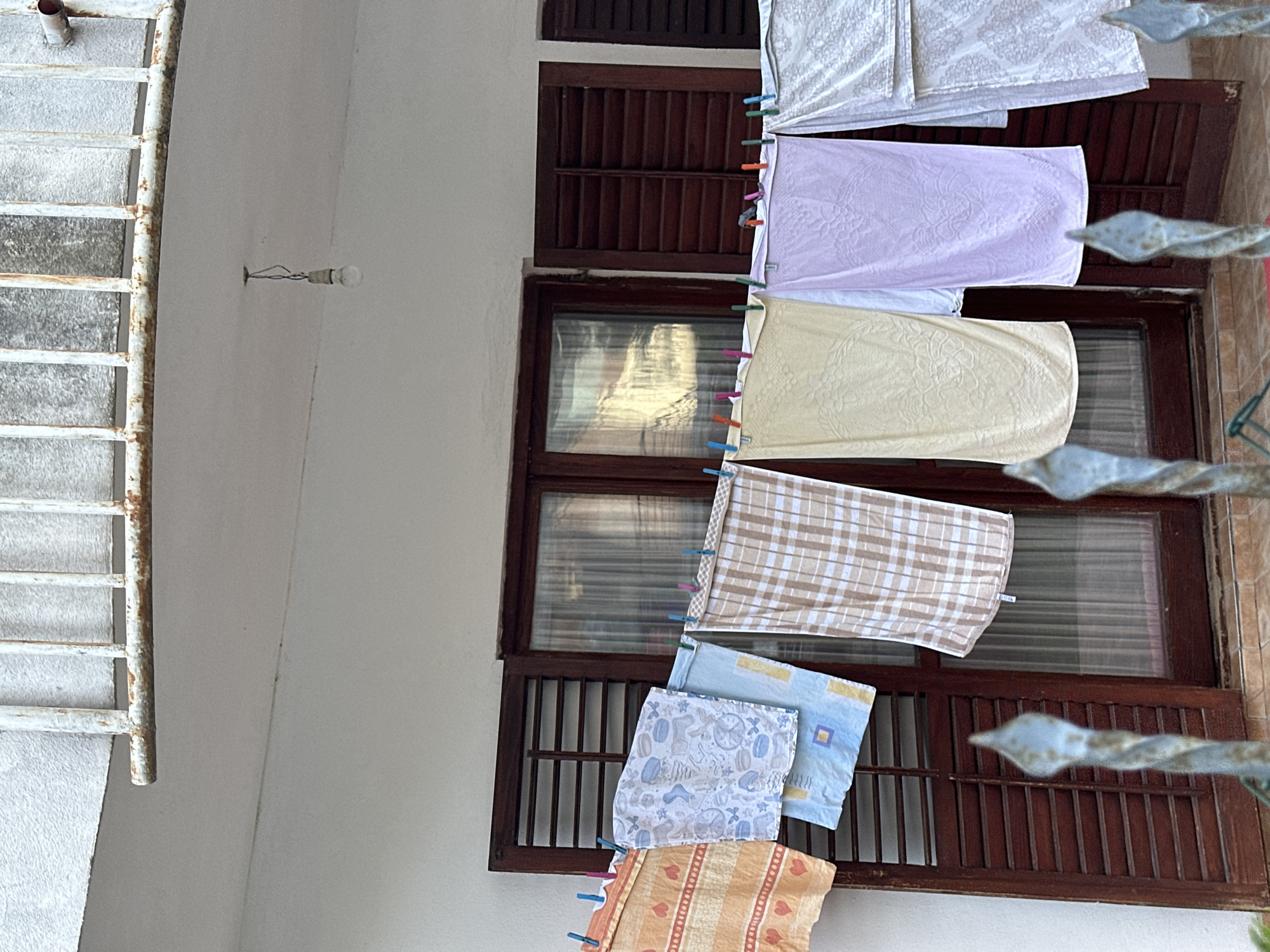 A clothesline with several towels hangs in front of a building with closed wooden shutters and a window. The towels are of various colors and patterns, including orange with hearts, blue with drawings, brown plaid, yellow, purple, and white. The scene appears to be outdoors, possibly in Budva, Montenegro, given the architectural style. A light bulb hangs from a wire overhead. In the foreground, there are parts of a decorative metal fence.