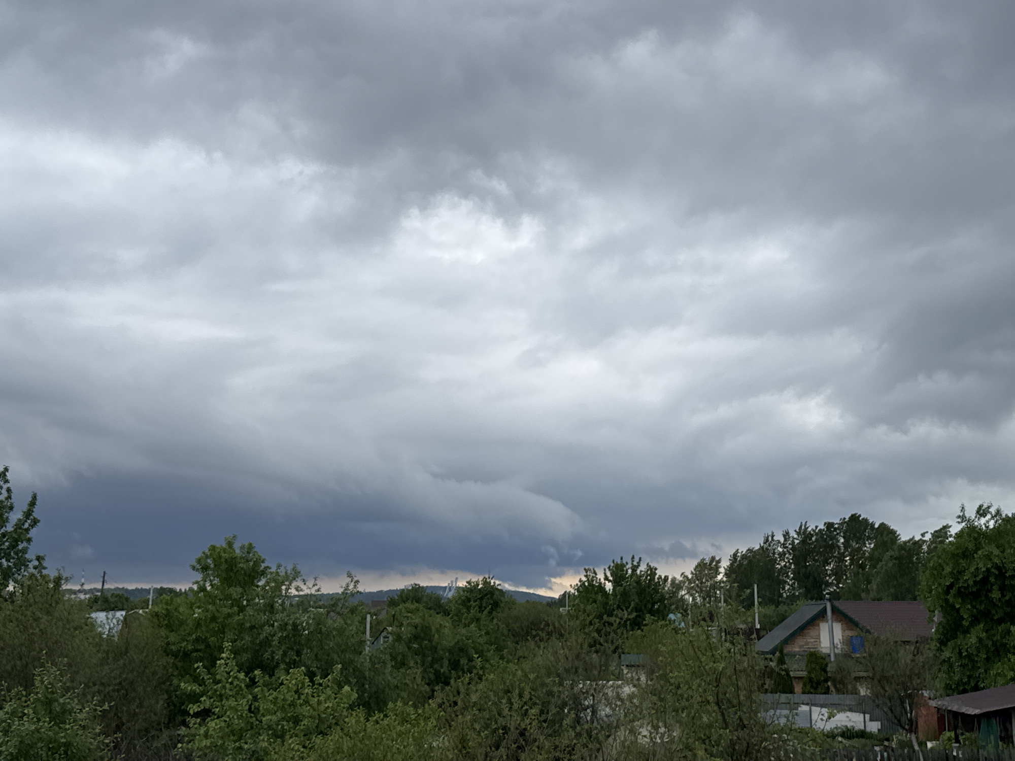 The image depicts an overcast sky over a suburban area, potentially within or near the city of Chaikovsky, Russia. The sky is dominated by dense, gray clouds, indicative of an impending storm or heavy rain. Below the cloud cover, a line of trees and vegetation fills the midground, obscuring much of the ground-level details. A few houses are partially visible among the trees, exhibiting typical residential architecture. In the distance, faint outlines of structures, possibly industrial or urban developments, can be seen against the horizon. There are no people visible in the image. The overall environment suggests a calm, rural setting under threatening weather conditions.