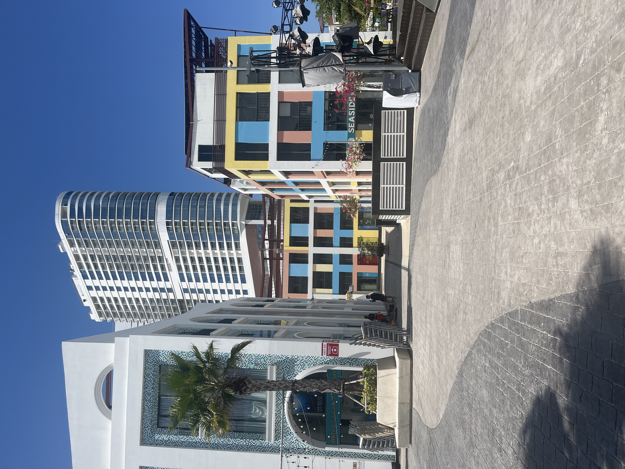 The image depicts a city street scene in an urban area, with a clear blue sky overhead. Three distinct buildings are visible. The first building is a white structure with an archway design, adorned with blue mosaic tiles and a palm tree. A sign with the words "AVATAR CINEMAS" is visible within the archway. To the left and behind the white building stands a modern, high-rise building with a curved facade and numerous windows. Adjacent to the white building is a colorful building with a grid-like design, featuring yellow, blue, peach, and white squares. The sign that says "SEASIDE" is visible on this building. On the right side of the image, a wide paved walkway is visible, with a distinctive curved pattern. A few individuals are present in the scene, appearing to be resting on a set of chairs in front of one of the buildings.