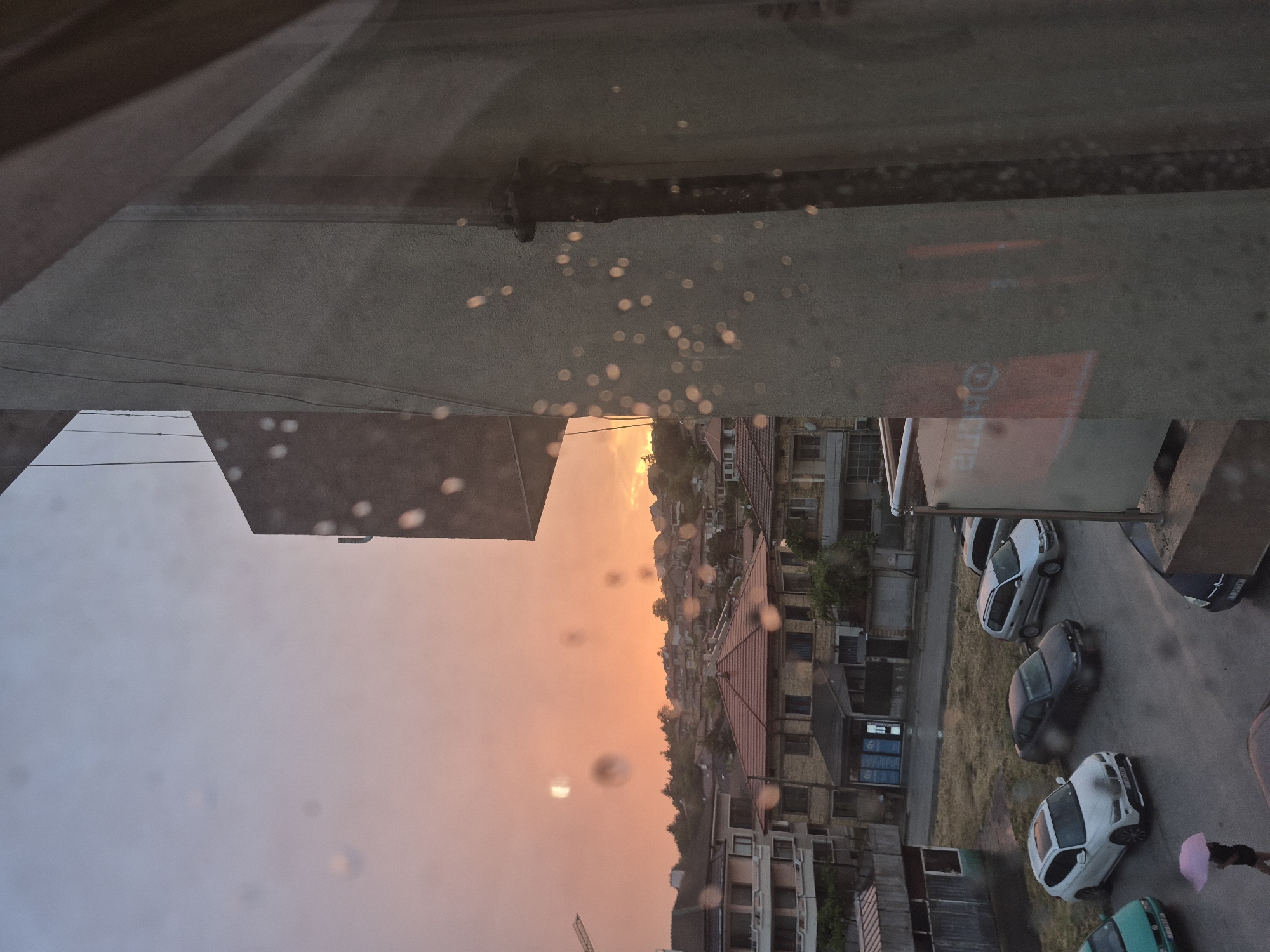 The image captures a view of the city of Popovo, Bulgaria, through a window with water droplets. The sky is a vibrant mix of orange and pink, indicating a sunset or sunrise. In the foreground, a concrete building structure is visible on the right, partially obscuring the view of the city. The city itself consists of residential buildings with red-tiled roofs and other structures. A parking lot is visible in the lower section of the image, with several cars parked, including a white SUV, a dark sedan, and a silver SUV. A person is walking in the parking lot holding a pink umbrella. The ground appears wet, suggesting recent rainfall. The overall atmosphere is overcast and moody, likely due to the weather conditions.