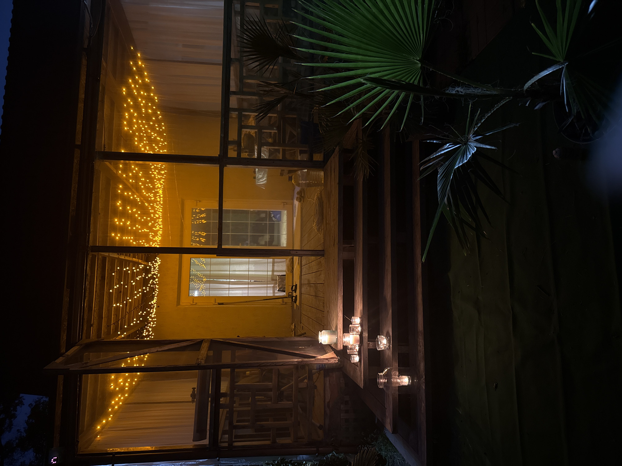 This is a nighttime, low-angle shot of an outdoor seating area with a wooden porch and stairs, illuminated by string lights and candles. The porch is covered by a roof, and the sides are partially enclosed by screens and white curtains. Several candles in glass holders are placed on the steps, creating a warm, inviting atmosphere. In the foreground, a palm tree with green fronds is visible. The area appears to be a private residence in Serhiivka, Ukraine. The overall mood is serene and peaceful.