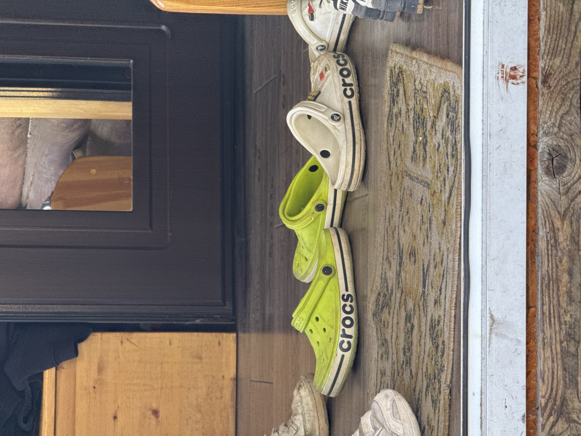 The image, taken in what appears to be an entryway or mudroom in Krivko, Russia, shows several pairs of shoes arranged on a floor next to a doorway. The scene is lit by indirect light, with no specific time of day discernible.

Dominating the center are three pairs of shoes. A pair of white Crocs is placed with its "CROCS" logo visible in black on the side strap. Above it, another white shoe, possibly a sneaker, features a partially visible "NIKE" logo. Below the white Crocs are a pair of bright lime green Crocs, also displaying the "CROCS" logo in black on the side strap. These green Crocs appear well-used. Further down, partially visible at the bottom of the frame, are a pair of dirty white sneakers.

The shoes rest on a light brown wooden floor. To the right of the shoes, a patterned rug with a muted yellow and dark abstract design is visible. Beyond the rug, there's a light-colored doorframe or trim, and further right, a weathered wooden surface, possibly a wall or door, with a prominent knot in the wood.

To the left, a dark brown or black door frame with a small rectangular glass panel is visible. The glass reflects a light-colored, textured object, possibly insulation or a light-colored wall inside. Below this door frame, a wooden structure or piece of furniture is partially seen. No people or active events are depicted; the scene suggests a casual collection of footwear near an entrance.