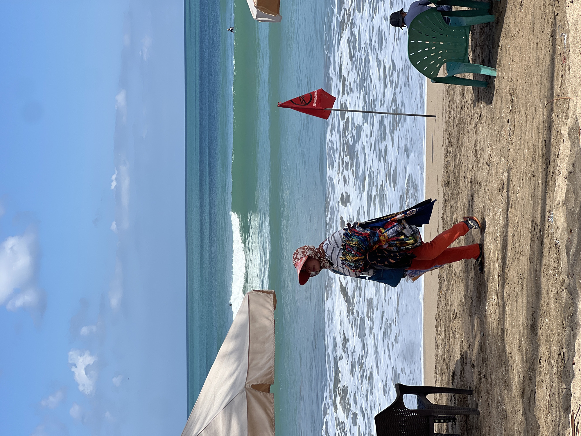 A woman walks along the sandy shore of Kuta Beach, Indonesia, on a bright, sunny day with a blue sky and scattered clouds. She wears an orange outfit and a headscarf, carrying a bundle of colorful textiles. The ocean is a vibrant turquoise with moderate waves, and a red flag with the word "SWIMMING" is planted in the sand near the water's edge. Beach chairs are visible on the sand, and a surfer is in the distance.