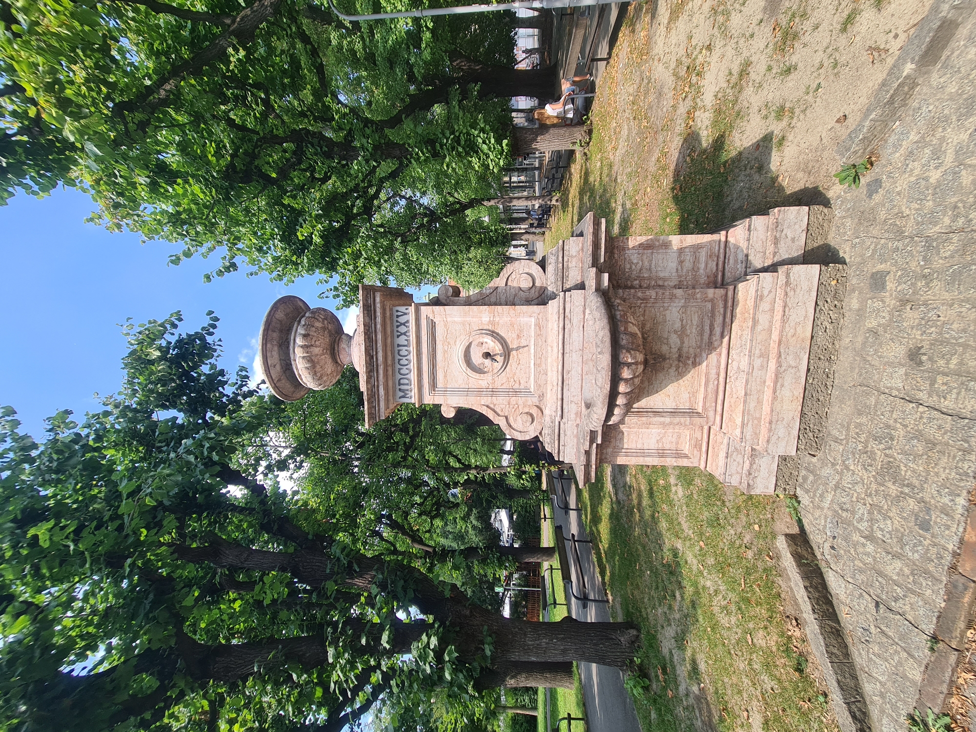 The image shows a pink marble fountain in a park in Vienna, Austria. The fountain has a large urn on top and a carved inscription that reads "MDCCCLXXV". In the background, there are trees, benches, and people sitting and walking. The sun is shining and the sky is blue. The park appears to be well-maintained and offers a peaceful setting. The fountain is the main subject of the image, with the surrounding park serving as a backdrop. The people are casually enjoying the park's amenities.